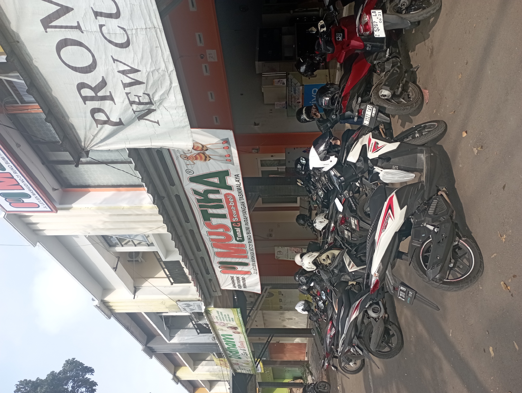 The image depicts a street scene in Tasikmalaya, Indonesia, likely in a commercial area. In the foreground, a row of parked motorcycles is visible, many with helmets placed on them. The motorcycles vary in color and model, suggesting a common mode of transportation.
Behind the motorcycles, a storefront with an orange awning extends across the width of the image. Above the awning, there are several signs. One prominent sign, partially obscured, reads "PROM NEW CU". Another sign, to the left, is for "Didham Spa & Beauty" and lists services such as "Haircuts, Blow Dryer, Creambath, Coloring, Hair Spa, Facial, Manicure, Pedicure, Waxing, Eyebrow, Massage, Body Treatment". Further to the left, a smaller sign is visible with text that is difficult to decipher.
Above the storefront, a multi-story building with windows and balconies is visible. The architecture suggests it is an older commercial building. The overall impression is a busy street in a city, with commerce and transportation being central elements of the scene. The lighting suggests it is daytime, with sunlight casting shadows on the ground.