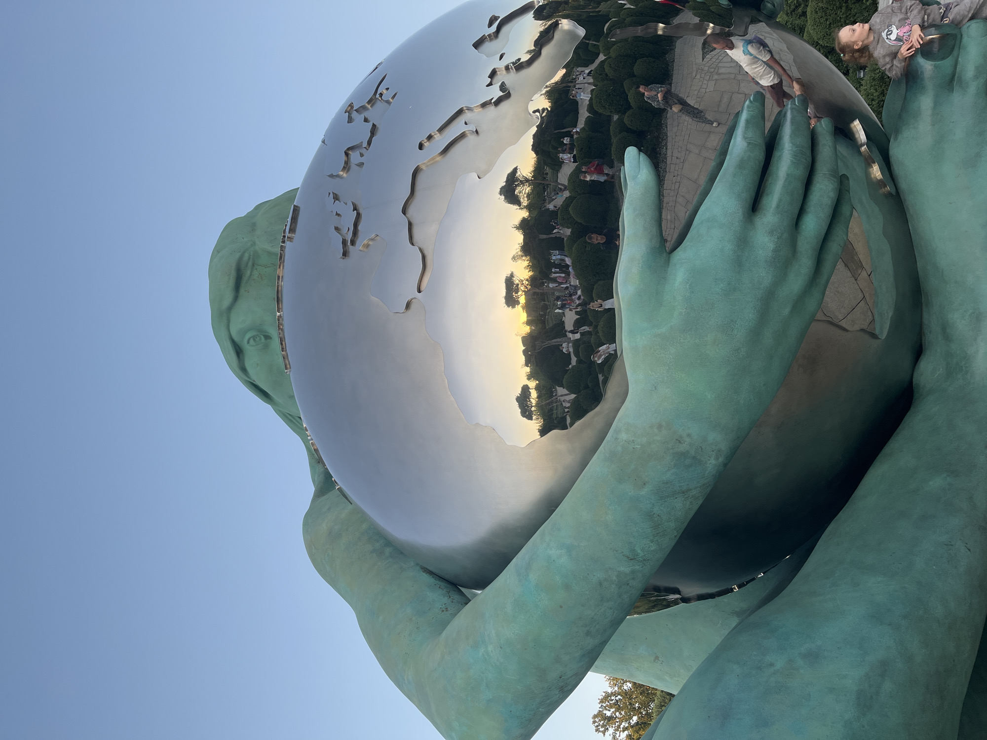 The image shows a large, abstract sculpture of a figure cradling a metallic globe, reflecting a park-like setting with people. The sculpture is made of a green material, possibly bronze, which has a weathered patina. The globe is highly polished and reflects the surrounding environment, including trees, bushes, pathways, and several individuals. The sky above is clear and blue, suggesting it is daytime. In the foreground, a young girl with curly hair, wearing a gray sweatshirt with a cartoon design and gray sweatpants, is looking towards the viewer and leaning on a railing. The sculpture is situated in a park, possibly the "Solnechny Ostrov" (Sun Island) Amusement Park in Krasnodar, Russia, given the style of the sculpture and its common depiction in tourist photos from that location. The reflection in the globe shows a sunset with trees and more people scattered throughout the park.
