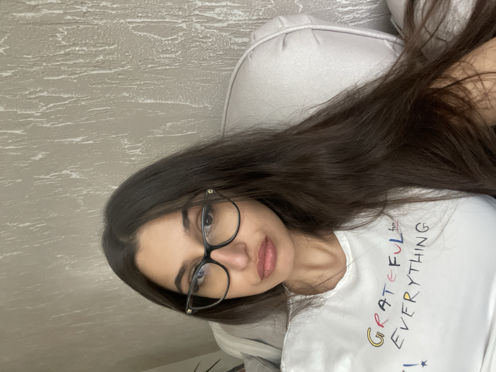 A woman with long, dark hair and glasses is lying on a gray, cushioned surface, possibly a sofa or bed, against a textured, light gray wall. She is wearing a white t-shirt with the word "GRATEFUL" printed on it in multi-colored letters, followed by "for EVERYTHING" below it. The location is likely indoors in Unknown, Unknown.