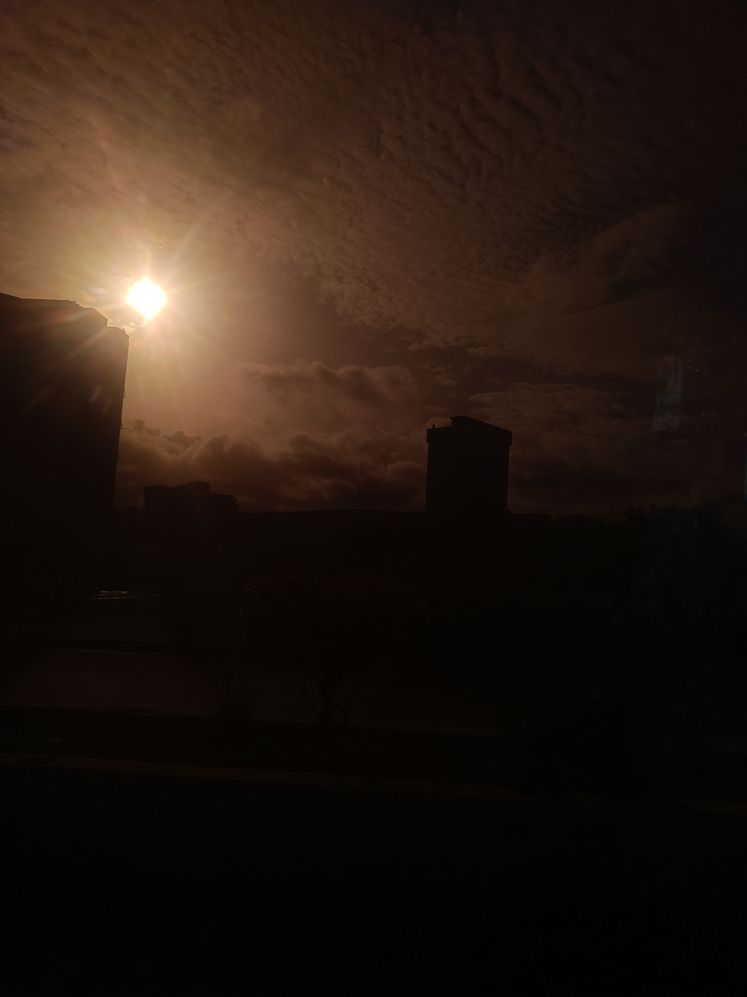 The image captures a sunset over Porlamar, Venezuela. The sun is bright and partially obscured by clouds, casting a warm, reddish-orange glow. Silhouettes of buildings, likely apartments or offices, are visible against the dramatic sky. The foreground is dark, with some indistinct shapes that could be trees or other structures. The overall impression is one of urban twilight, with the city bathed in the fading light of day.