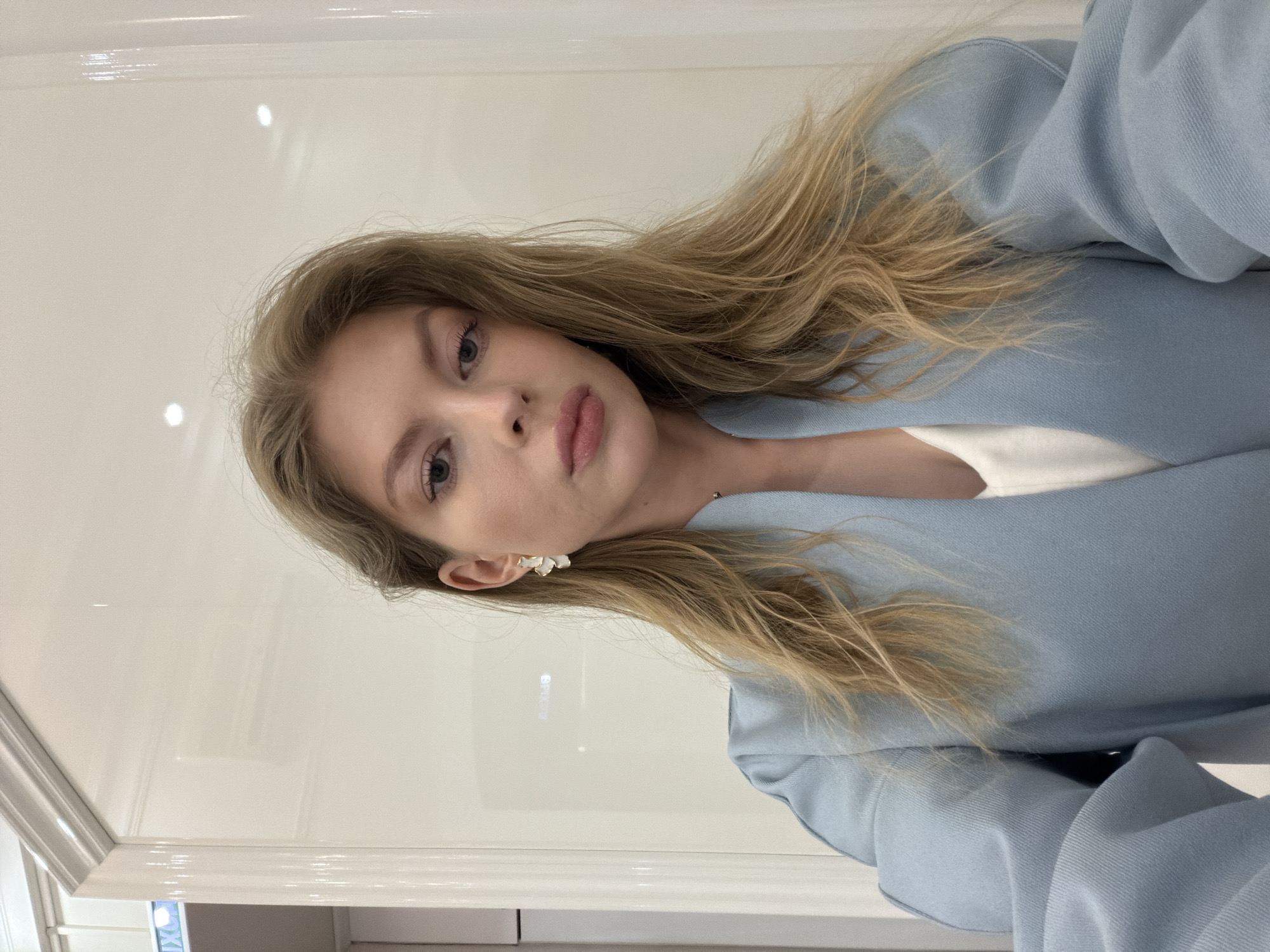 A blonde woman in a light blue jacket poses indoors. The background consists of a glossy white wall, suggesting a modern interior. The location cannot be determined.