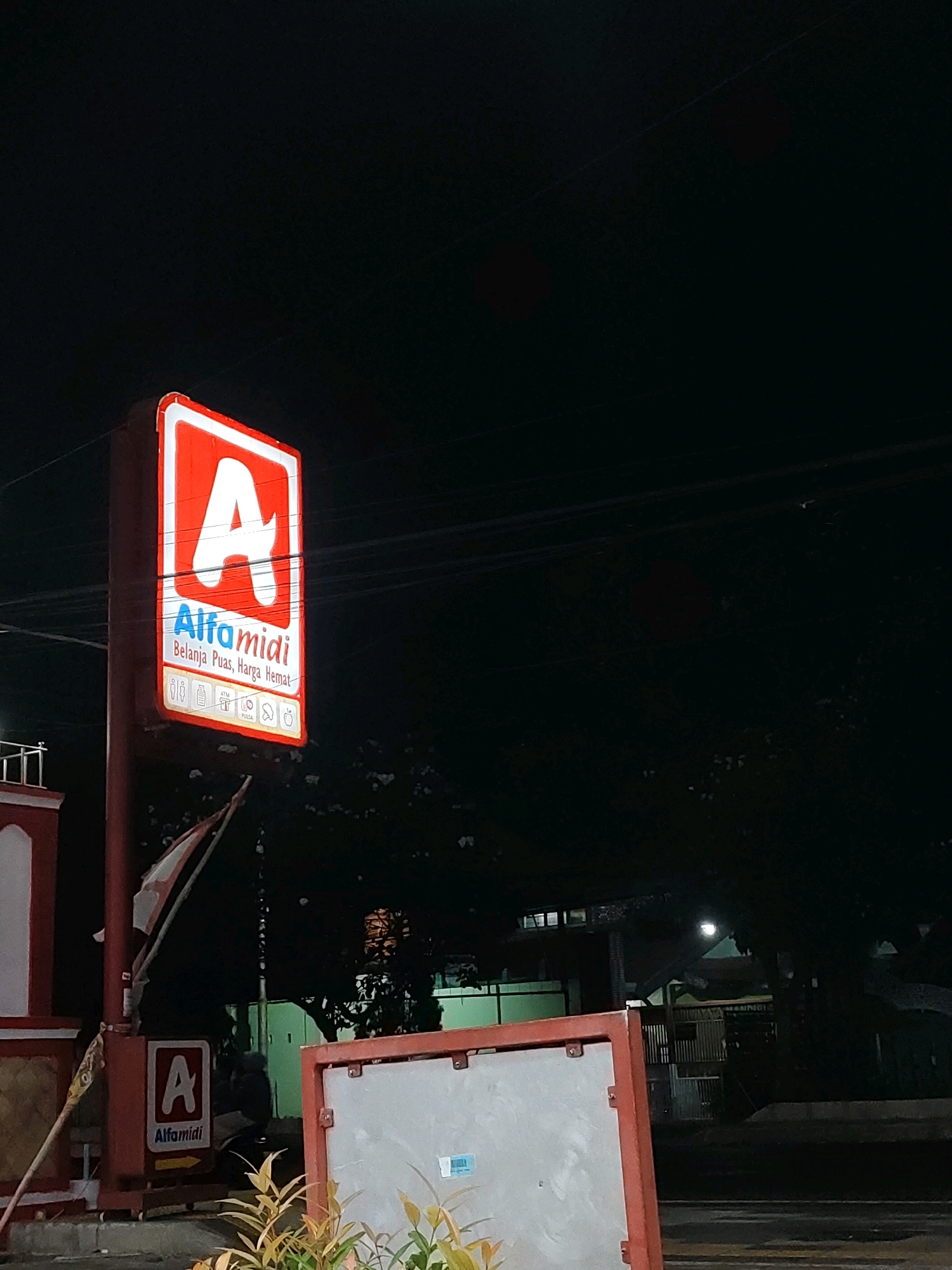 The image shows a night scene in Probolinggo, Indonesia, with a prominent, illuminated "Alfamidi" store sign as the central subject. The sign is red and white, featuring a large "A" logo above the store name in blue lettering. Below the store name, it reads "Belanja Puas, Harga Hemat" (Satisfying Shopping, Affordable Price), with several small icons representing services like ATMs and restrooms. The sign is mounted on a red pole.
To the left of the main sign, a smaller "Alfamidi" sign with an arrow pointing right is visible, attached to a red structure that appears to be part of the store's facade, which is also red and white. A partial view of a building with a white column and a red base is present.
In the background, dark trees create a silhouette against the night sky. Some faint lights are visible within the trees and in the distance, suggesting other buildings or streetlights. Power lines crisscross the upper portion of the image. A portion of a red-framed board is in the foreground, with a light-colored surface and a blue sticker. The ground in the foreground appears to be a wet road or sidewalk. No people are clearly visible in the image, though a shadow of a person might be discerned near the smaller Alfamidi sign.