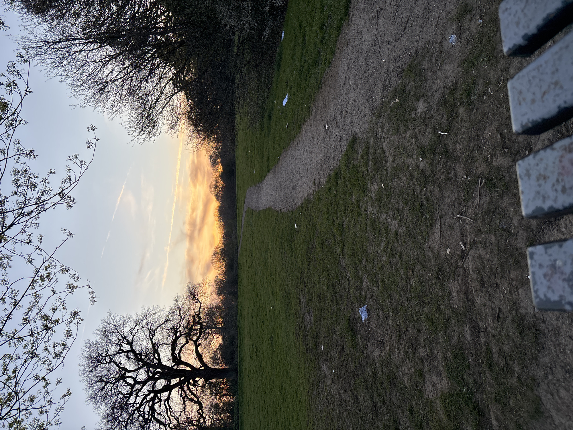 The image depicts a path in a park during sunset, located in Broxbourne, United Kingdom. The path extends diagonally from the lower right to the upper left of the frame, appearing to be made of dirt or gravel. Grass surrounds the path, with litter scattered across the grass and the path itself. Trees are visible on both sides of the path, silhouetted against the sky. The sky is a mix of pale blue and orange hues due to the setting sun. The edge of a metal bench is visible on the right side of the image.
