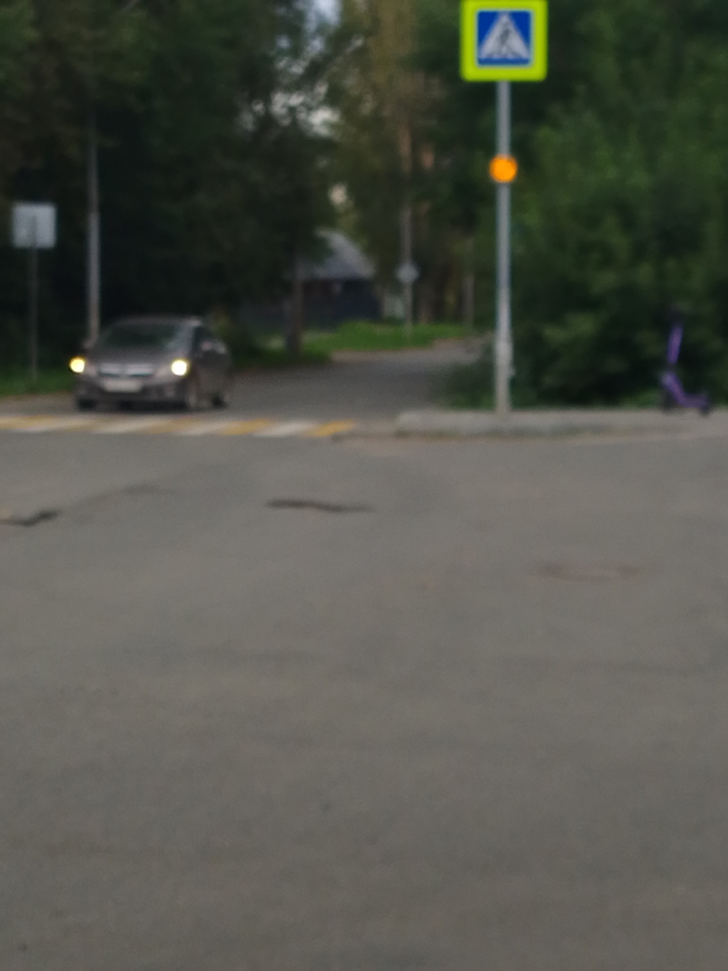 In the city of Unknown, Unknown, a gray sedan with its headlights on is stopped at a pedestrian crossing. The crossing is marked with yellow and white stripes. A blue and white pedestrian crossing sign with a yellow border is visible to the right of the car, mounted on a pole. An orange circular light is also attached to the same pole. Beyond the pedestrian crossing, trees and a building are visible. A purple electric scooter is parked on the right side of the image, behind a concrete curb. The scene appears to be an outdoor road setting, possibly in the evening or on an overcast day, given the car's headlights are on and the lighting is dim.