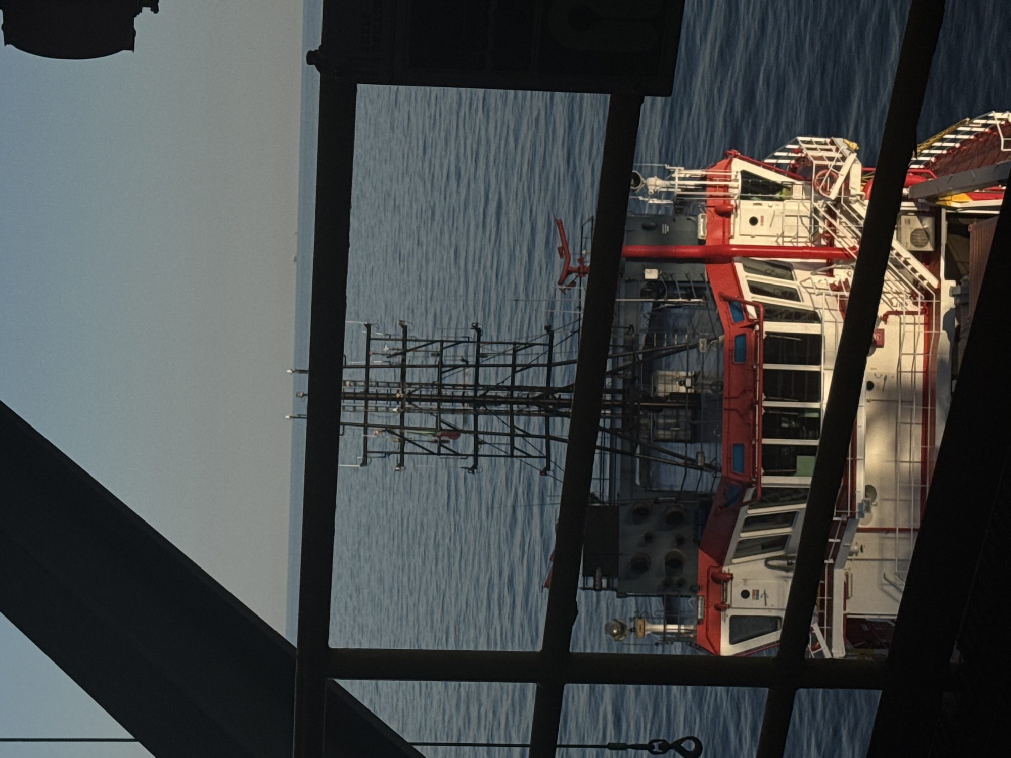 An eye-level, slightly angled, medium shot captures the stern of a red and white vessel at sea, seen through a dark metal frame. The vessel is oriented towards the viewer and appears to be a tugboat, with a prominent superstructure featuring multiple windows and a red roof. A tall, intricate metal mast with various antennas and lights rises from the deck. The vessel floats on calm, blue water, with a hazy horizon in the distance. The foreground is dominated by dark, abstract metal structures, suggesting the viewer is on another ship or platform. The sky above is a pale blue, transitioning to a lighter hue near the horizon. There are no discernible people or specific actions occurring. The location is unknown, and the image does not provide any contextual information about a city in Italy.