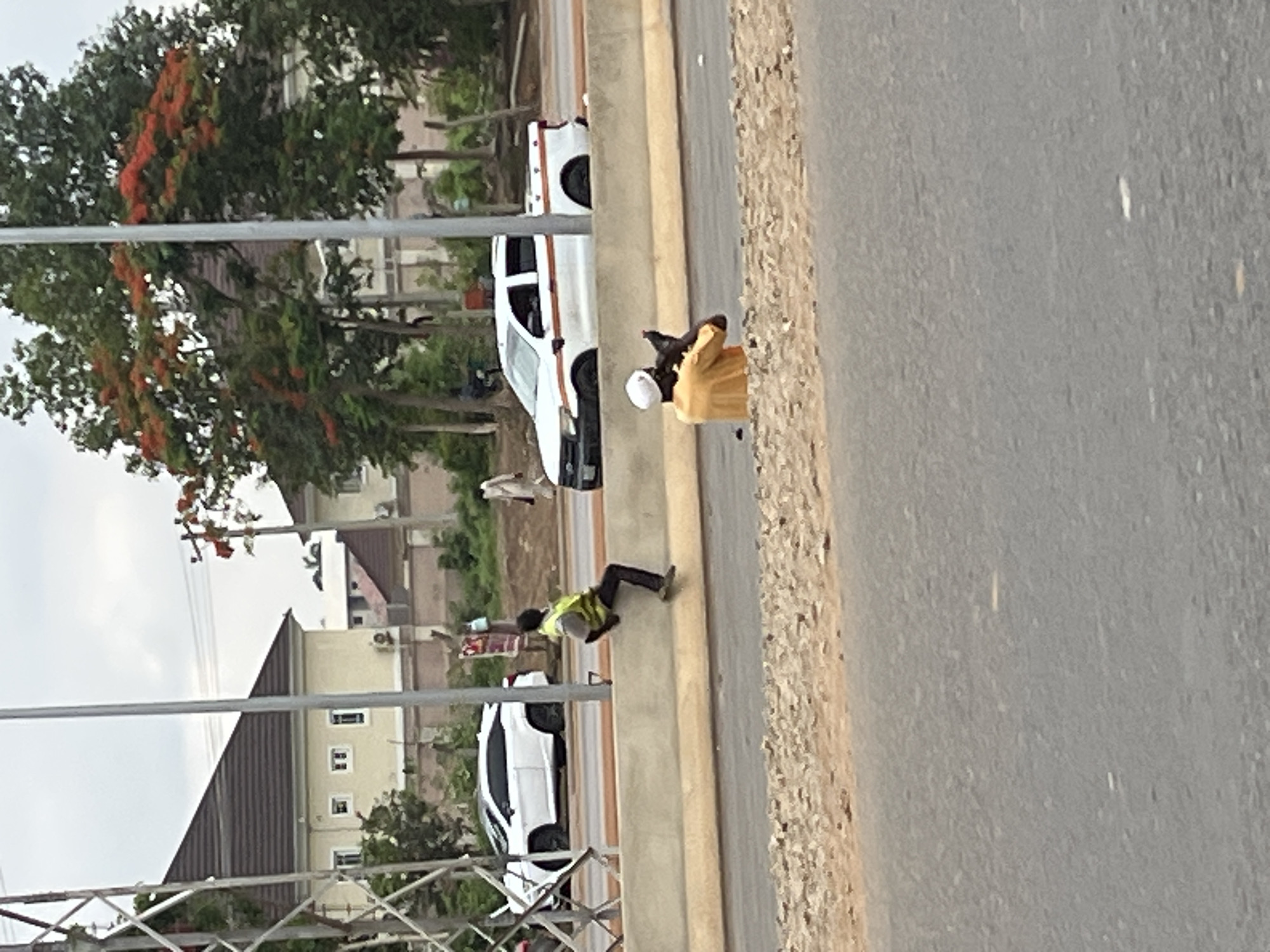The image shows a roadside scene in Abuja, Nigeria. Two white vehicles are parked along the side of the road next to a concrete barrier. Two individuals are present. One individual is wearing a yellow shirt and a white hard hat, and appears to be holding a bird, possibly a pigeon. Another individual in a fluorescent green vest is bending over the barrier. The roadside is separated from the road by a concrete barrier. A paved road stretches in the foreground, while buildings and trees are visible in the background. An electrical infrastructure is visible in the foreground.