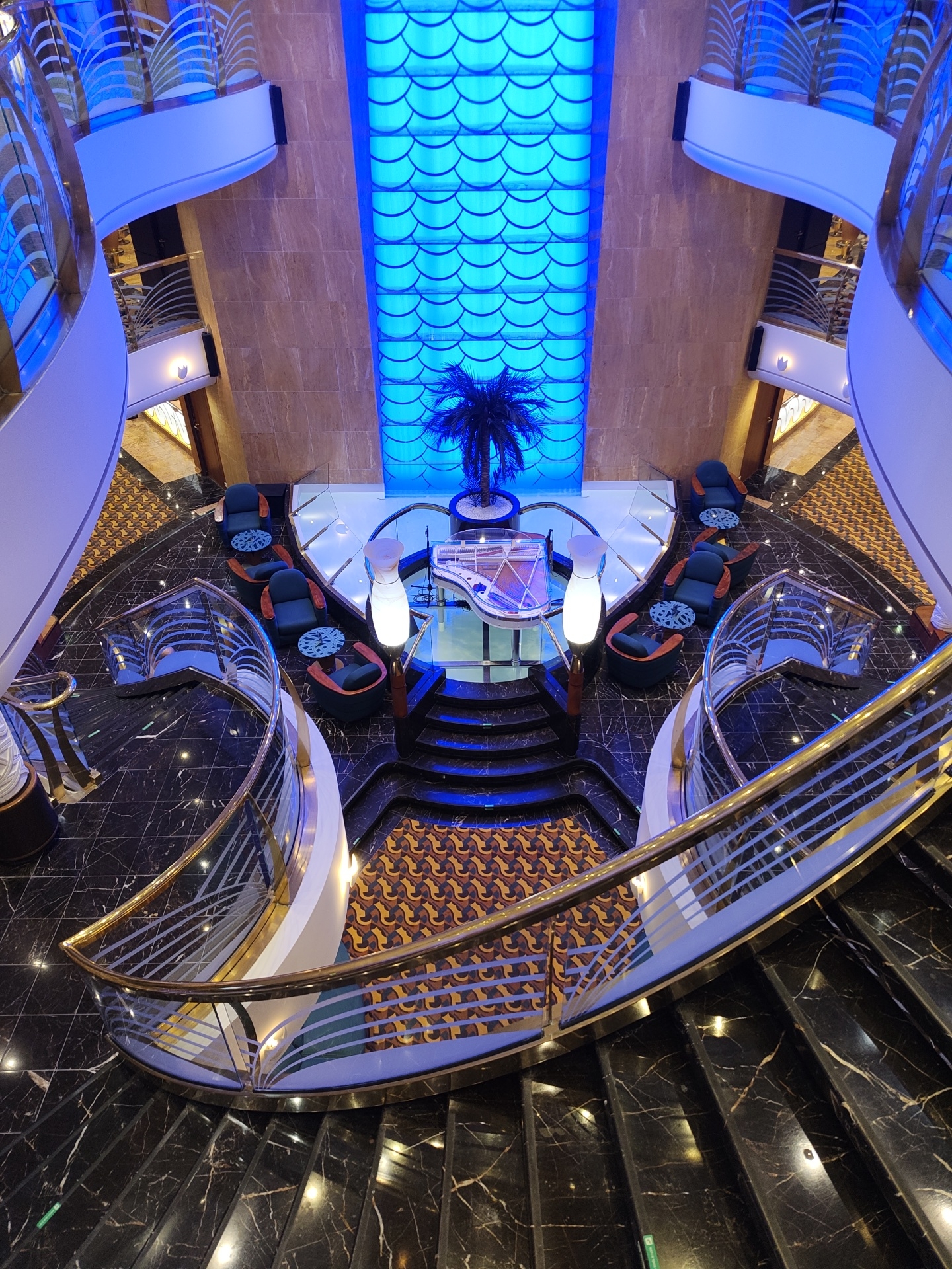 The image displays an opulent, multi-level interior space, likely a grand atrium or lounge within a luxury cruise ship, hotel, or upscale building. No people are visible.
The central area on the ground floor features a seating arrangement with dark teal armchairs and small, circular tables with intricate patterns. A white grand piano rests on a raised, light-colored platform, flanked by two tall, glowing white cylindrical lamps. Behind this central setup, a small potted palm tree stands against a striking back wall panel that is brightly illuminated with blue light, showcasing a repetitive scale-like or scallop shell pattern.
The floor is predominantly black marble with distinct white veining, complemented by a geometric patterned carpet in shades of brown, gold, and black underneath the seating area. Elegant, curving staircases made of the same black marble ascend from the ground level, leading to upper balconies. These staircases and balconies are outfitted with luxurious railings featuring gold-toned handrails, clear glass panels, and frosted horizontal accents, all dramatically lit with blue light, creating a sophisticated and somewhat ethereal ambiance. The upper walls appear to be clad in a light brown marble or stone, providing a warm contrast to the predominant cool blue lighting. The scene evokes a sense of modern luxury and is set up for relaxation, social gatherings, or live entertainment. No visible text is present.