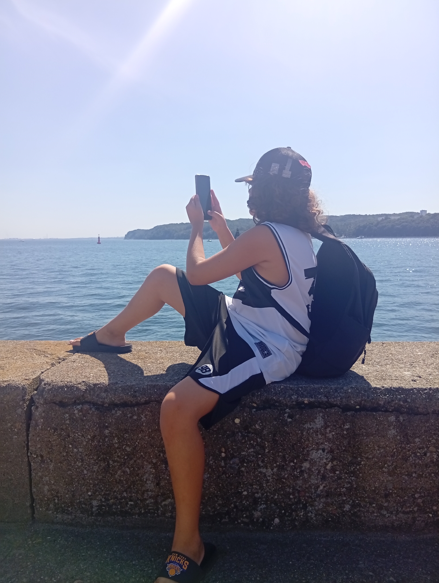 A person is sitting on a concrete barrier overlooking the sea. They are wearing a black baseball cap, a white tank top with black trim, black shorts with white trim, and black slides. A black backpack is resting behind them. The person is holding a smartphone and appears to be taking a picture or video. The sea is a vibrant blue, and a tree-covered hillside is visible in the distance. The sun is shining brightly, casting a lens flare across the sky. There is a red buoy in the water. The location is likely the area around the Gdynia Aquarium or the southern part of the city's waterfront, given the characteristic coastal scenery and urban development in the background.