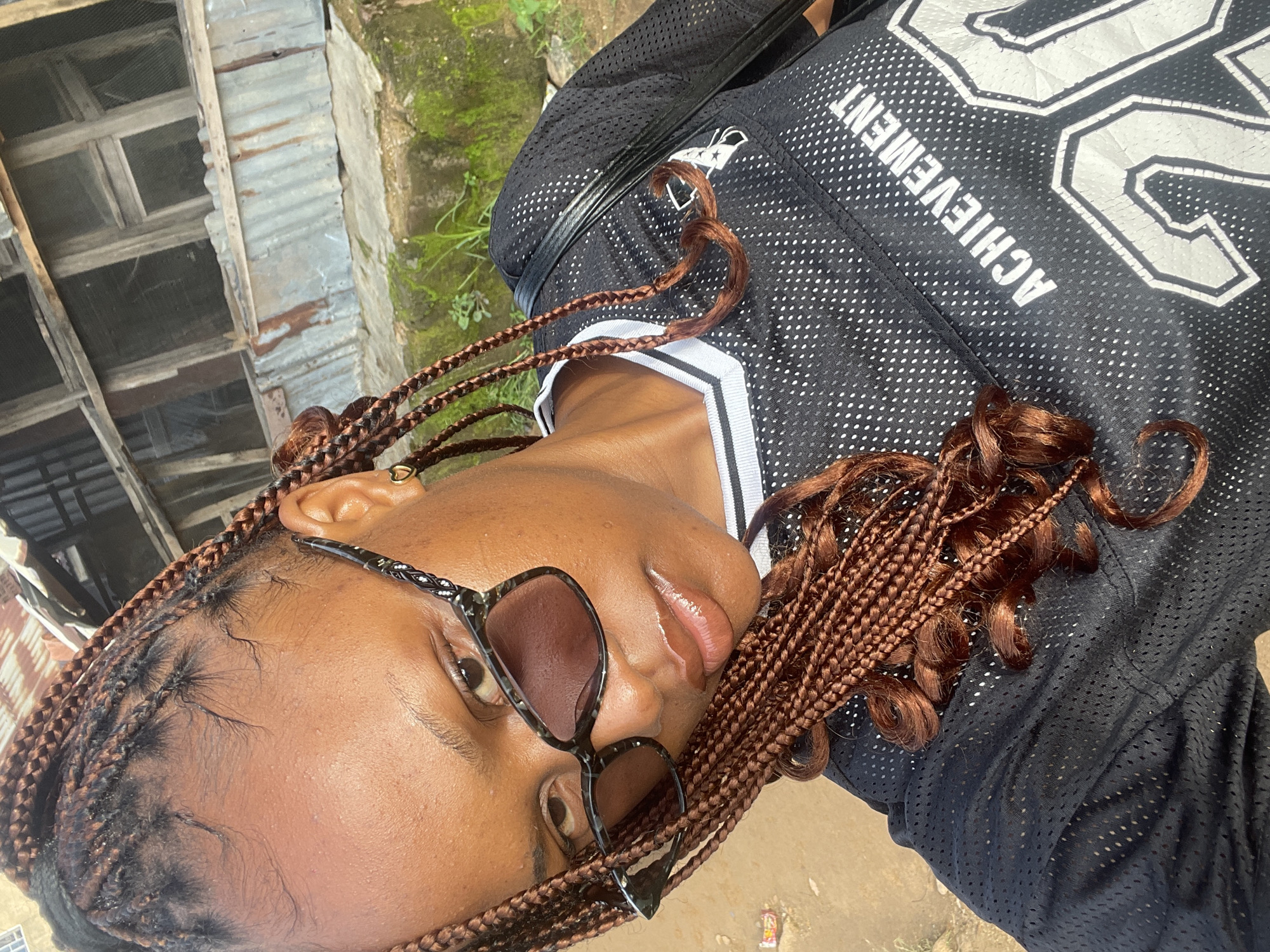 A young Nigerian woman with brown braided hair and sunglasses is posing for a selfie outdoors. She is wearing a black athletic jersey with the word "ACHIEVEMENT" and the number "20" visible. The background suggests a semi-urban or rural setting, possibly in Rafin Par, Nigeria, with a rustic wooden structure and corrugated metal sheeting visible, along with some overgrowth of greenery and dirt ground. The lighting indicates it is daytime.