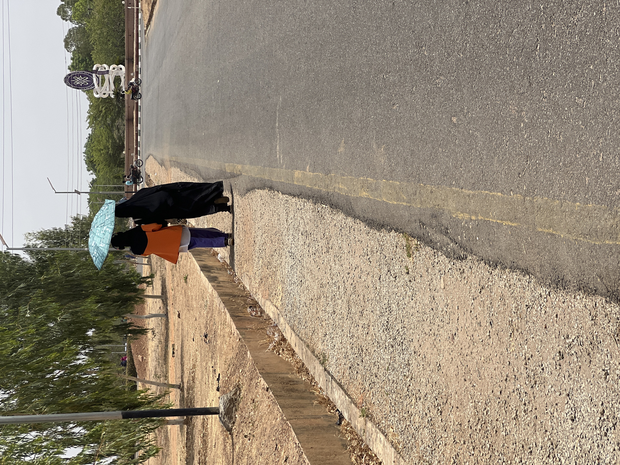 The image depicts two individuals walking away from the camera on a gravel shoulder adjacent to an asphalt road. This scene is located within the Federal University Gusau campus in Gusau, Zamfara State, Nigeria, as indicated by a prominent blue and white circular emblem with the inscription "FEDERAL UNIVERSITY GUSUA" on a sculpture in the background.
The person on the left wears an orange jacket over a lighter garment, dark trousers, and a dark head covering, holding an open light blue umbrella to shield both individuals. The person on the right is dressed in a full-length black garment. They are walking in close proximity. To the left of the gravel shoulder, a concrete drainage ditch runs parallel to the road, containing dry leaves and debris. Further left, a line of trees with long, slender green leaves stands beside a dark gray lamp post. The asphalt road features a faded yellow line marking the edge of the paved surface. In the middle ground, on the main road, two individuals are visible riding separate motorbikes. The sky is bright and appears hazy or overcast. The immediate ground cover consists of dry, reddish-brown earth and gravel.