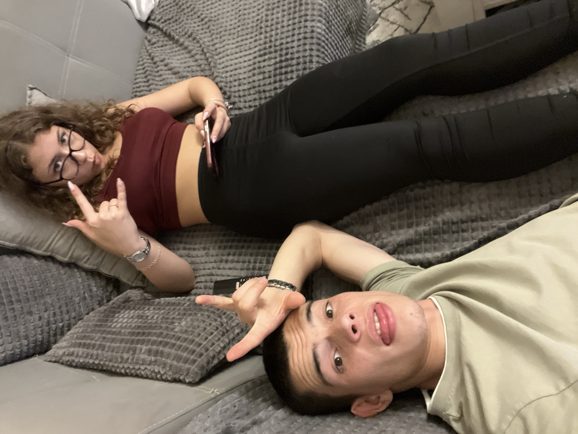 Two people are on a couch covered with a gray patterned blanket. A woman with curly hair and glasses is lying on her side, facing the camera and making a hand gesture. She is wearing a maroon crop top and black leggings. She holds a smartphone. A man is lying perpendicular to her at the bottom of the frame, with his head close to the camera and also making a hand gesture. He is wearing a tan shirt. The setting appears to be a living room. There is no visual evidence to determine if this image was taken in Plovdiv, Bulgaria.
