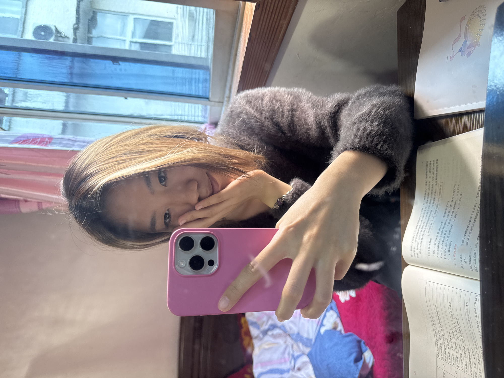 A young woman is taking a selfie indoors. She is holding a pink smartphone in her right hand, the camera pointing at her face. Her left hand is near her face, perhaps posing. She has shoulder-length blonde-streaked hair and is wearing a fuzzy grey sweater. Beside her is an open book with Chinese text. A window with a view of buildings, including an air conditioning unit, is in the background. The location is likely Hechuan, China, based on the text language in the book. The backdrop includes a bed with patterned bedding.