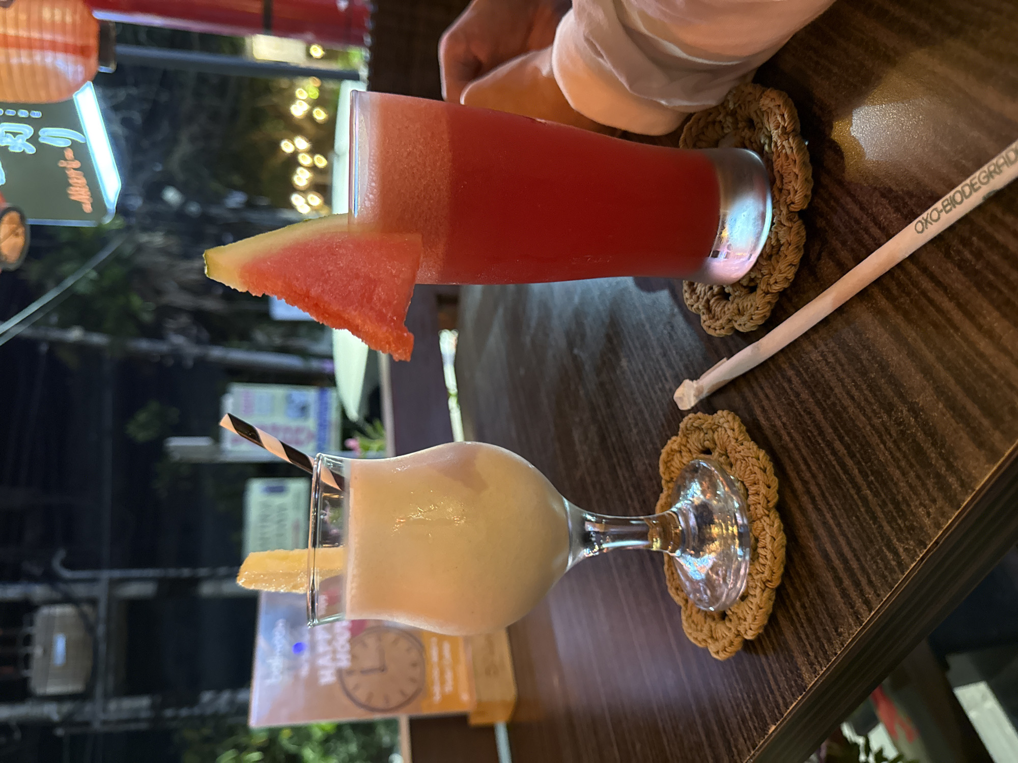 Two drinks are on a wooden table at a restaurant in Denpasar, Indonesia. The drink on the left is in a stemmed glass and appears to be a creamy, pale yellow smoothie or milkshake, garnished with a slice of fruit. The drink on the right is in a tall glass and is a vibrant red, likely a watermelon or strawberry juice, garnished with a wedge of watermelon. Both drinks are on woven coasters. A striped straw is in the left drink, and a paper straw lies on the table next to the right drink, with the visible text "OXO-BIODEGRADABLE" printed on it. In the background, there are blurred lights, foliage, and signage that appears to be in Japanese. A person's arm, wearing a white sleeve, is visible on the right side of the image.
