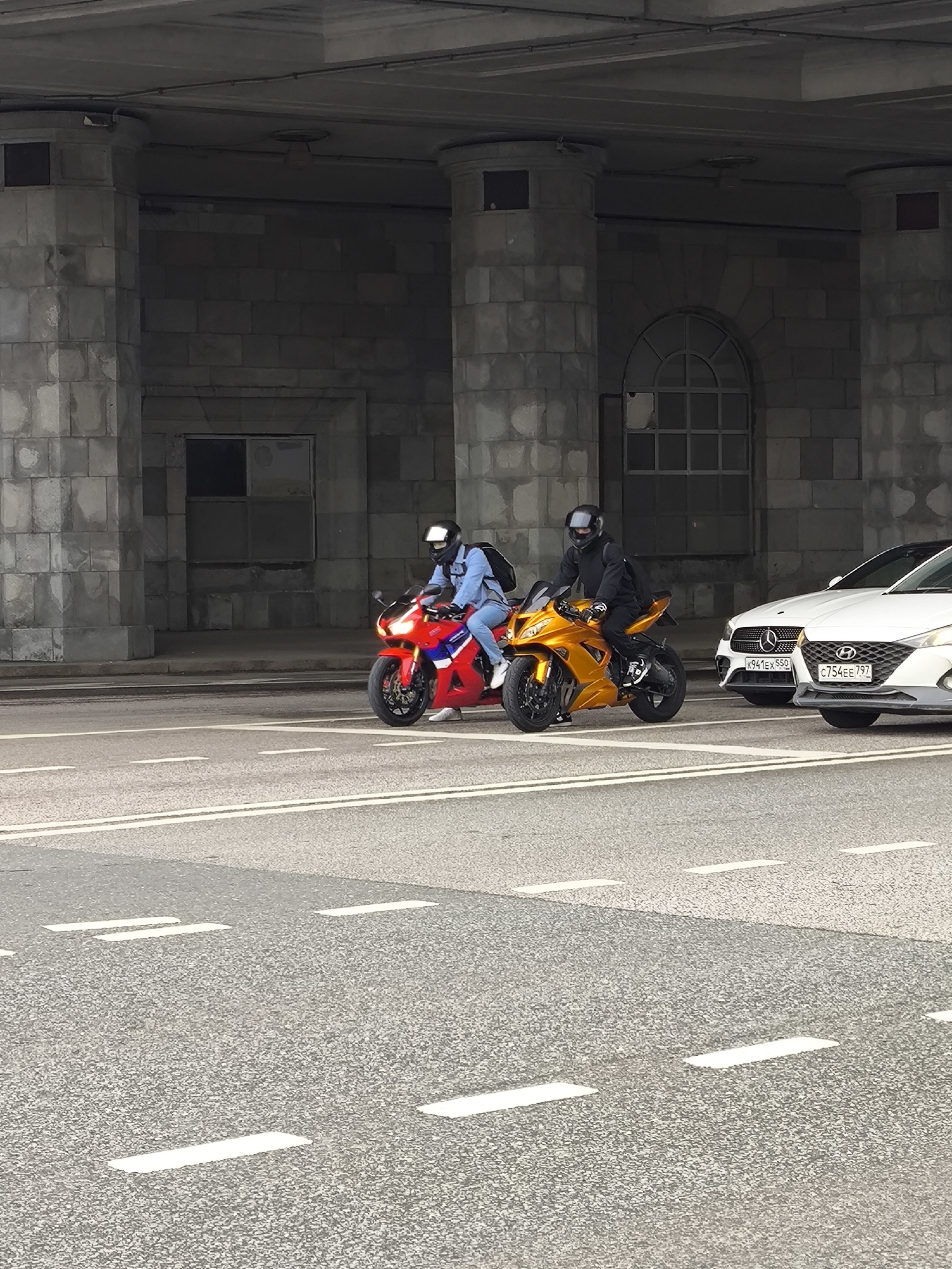 The image depicts a street scene in Moscow, Russia, with two motorcyclists on a road under a concrete overpass or bridge. The motorcyclists are riding a red and a gold-colored sport bike. The motorcyclist on the red bike is wearing a light blue jacket and a white helmet, while the other is wearing all black. To the right, partially visible, is a white car. The street has painted white lines marking lanes. The overpass/bridge is constructed of concrete blocks. The license plates on the white car are "K941EX550" and "C754EE797".
