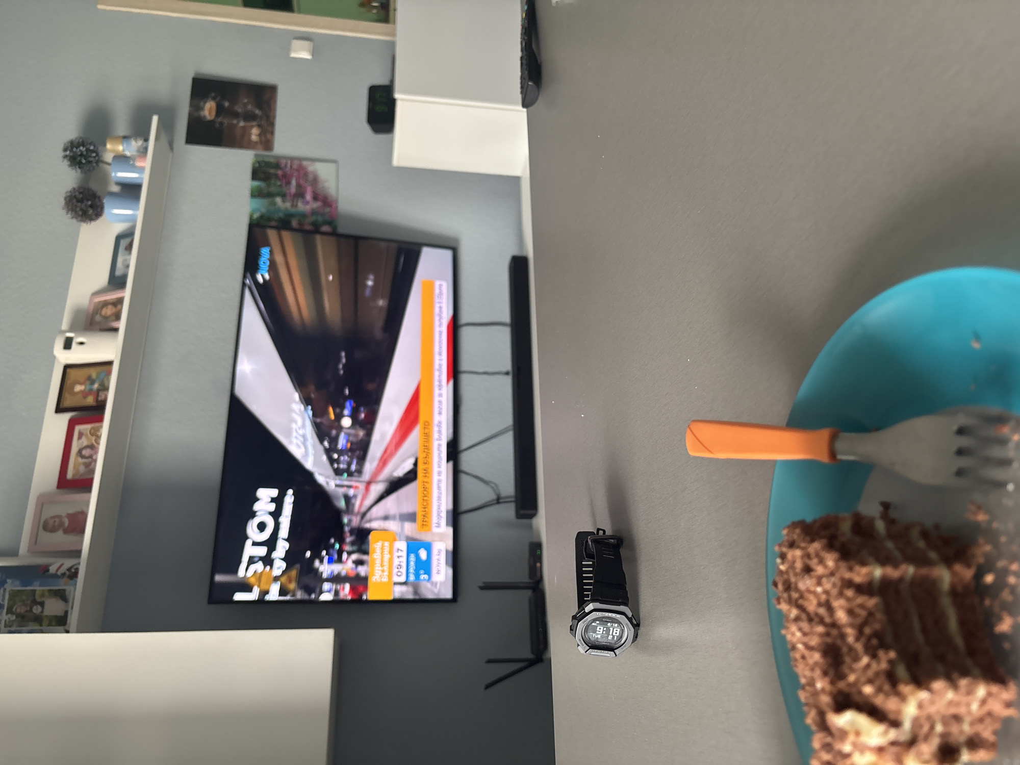 A close-up interior shot depicts a scene featuring a piece of chocolate cake on a blue plate with a fork, next to a black G-Shock watch displaying the time 9:18 AM and the date Tuesday, 6/18. In the background, a television displays a news program with the word "LSTOM" in a large, vertically-oriented font, along with Bulgarian text and "NOVA" in the upper right corner. The program appears to be reporting on transportation. The television screen also features text reading "Здравей Балкария", "Транспорт на Бъдещето". A shelf of photos is on the left side of the shot. A wall-mounted soundbar is near the television. The location is Sofia, Bulgaria, as "България" is visible on the TV screen.