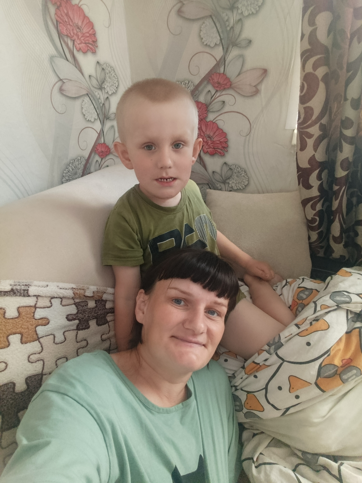 The photograph captures a woman and a young boy in an indoor setting. The woman, positioned in the foreground, has fair skin, short dark hair with bangs, and is wearing a light green t-shirt. She is smiling slightly. The boy, who appears to be sitting behind her, has fair skin, light blonde hair, and is wearing a green t-shirt with a dark graphic. He is also smiling.
The background features a wall with floral wallpaper, along with curtains with a brown and white pattern. The subjects are sitting or lying on a bed, with patterned bedding and pillows visible. The image is cropped closely around the subjects, obscuring the wider environment.
There is no evidence to identify the location as Фоки, Russia.