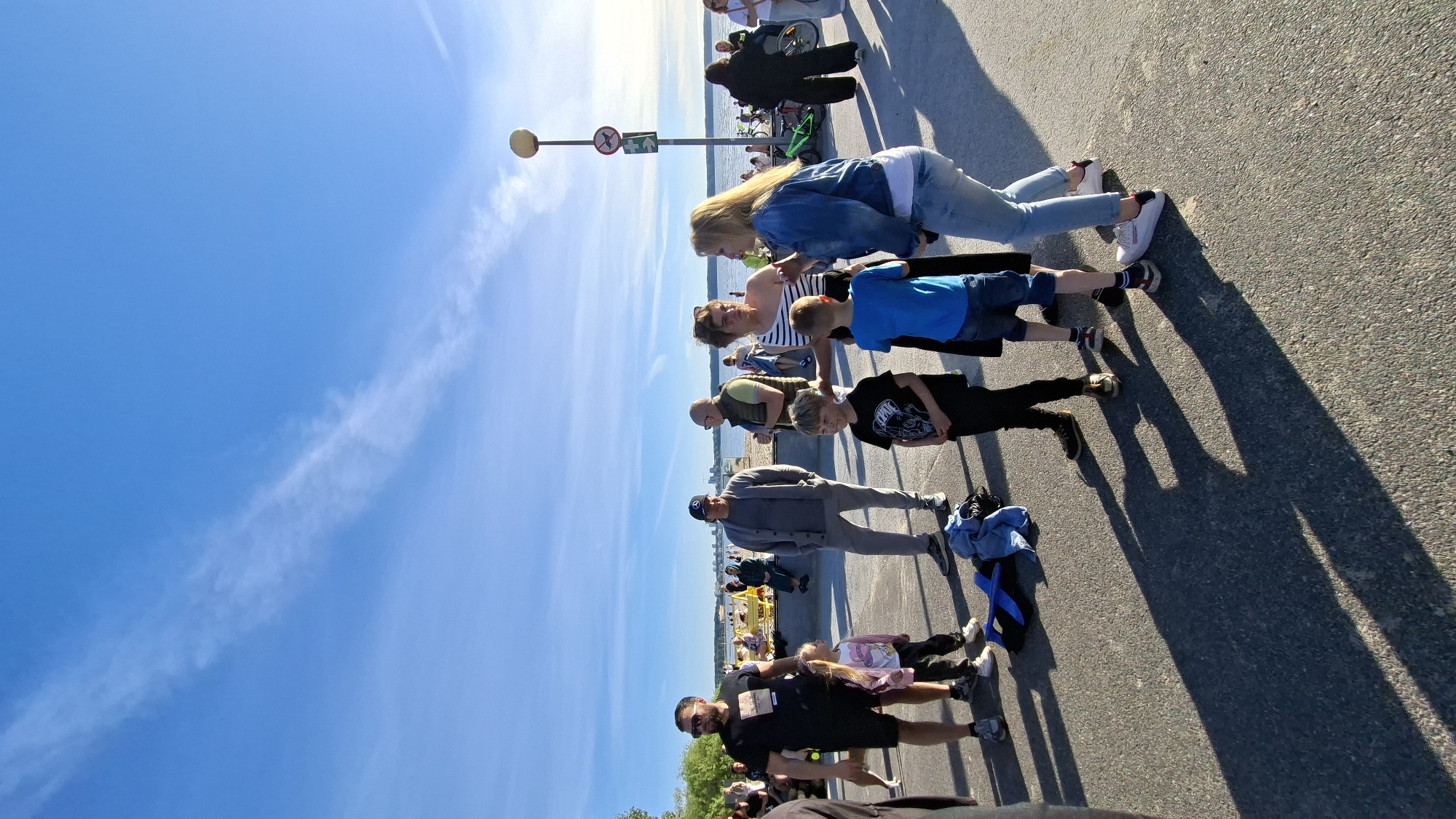 On a sunny day, a group of people are gathered on a paved area near a beach. The sky is blue with some wispy clouds. In the background, a body of water is visible, with a city skyline in the distance. Several people, including adults and children, are standing and talking. Some are dressed casually in t-shirts, shorts, and jeans. One man is wearing a black t-shirt and shorts, while a woman is in a denim jacket and jeans. Children are also present, one in a black t-shirt and another in a blue t-shirt. There is a street lamp with signage and a trash bin nearby. The ground is asphalt, with long shadows cast by the people and objects. A few bicycles are visible in the background, and some people are sitting on a low wall or bench near the beach. The scene appears to be a public outdoor space, possibly a waterfront promenade. The presence of the city skyline in the distance suggests this is an urban or suburban setting.