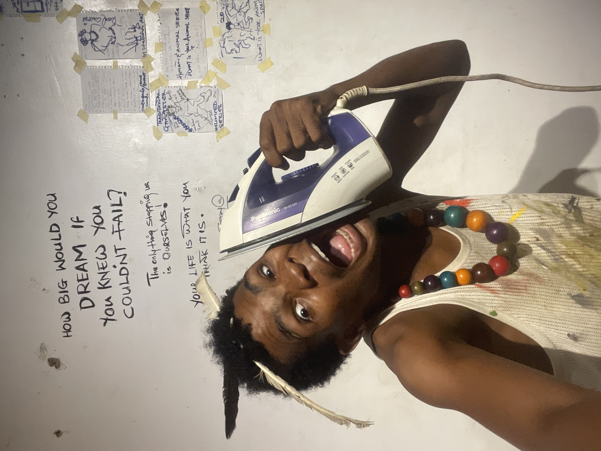 A young Black person with feathers in their hair holds a white and purple iron to their ear against a white wall. They are wearing a white tank top with colorful paint splatters and a beaded necklace. The wall has several handwritten messages and taped-up drawings. The handwritten messages include: "HOW BIG WOULD YOU DREAM IF YOU KNEW YOU COULDN'T FAIL?", "The only thing stopping us is OURSELVES!", and "YOUR LIFE IS WHAT YOU THINK IT IS." The taped-up drawings include a figure labeled "TRADITIONAL GANGSTER UNLIMITED SERIES" and two drawings with the question "WHAT IS YOUR ANIMAL SPIRIT?". The scene appears to be an indoor studio space in Ado, Nigeria.