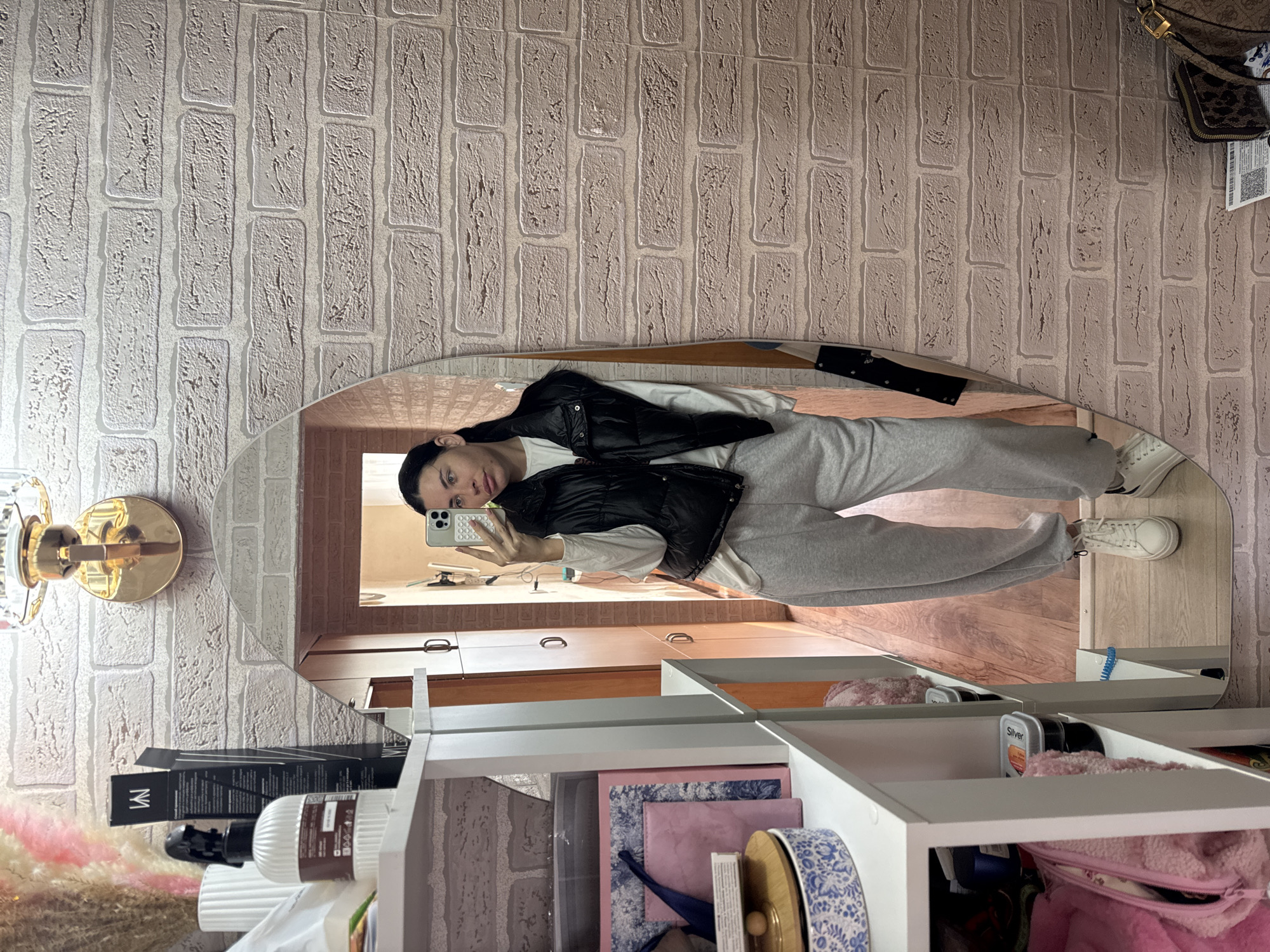 In a room with a brick-patterned wallpaper, a young woman stands in front of a full-length oval mirror, taking a selfie with her phone. She is wearing a white long-sleeved shirt, a black puffer vest, gray sweatpants, and white sneakers. To her left, a white shelving unit holds various items, including boxes, bottles, and bags. A wooden floor is visible in the background, and a doorway leads to another part of the room. The overall impression is a casual indoor setting, possibly a bedroom or dressing area. The location is specified as Chaikovsky, Russia.