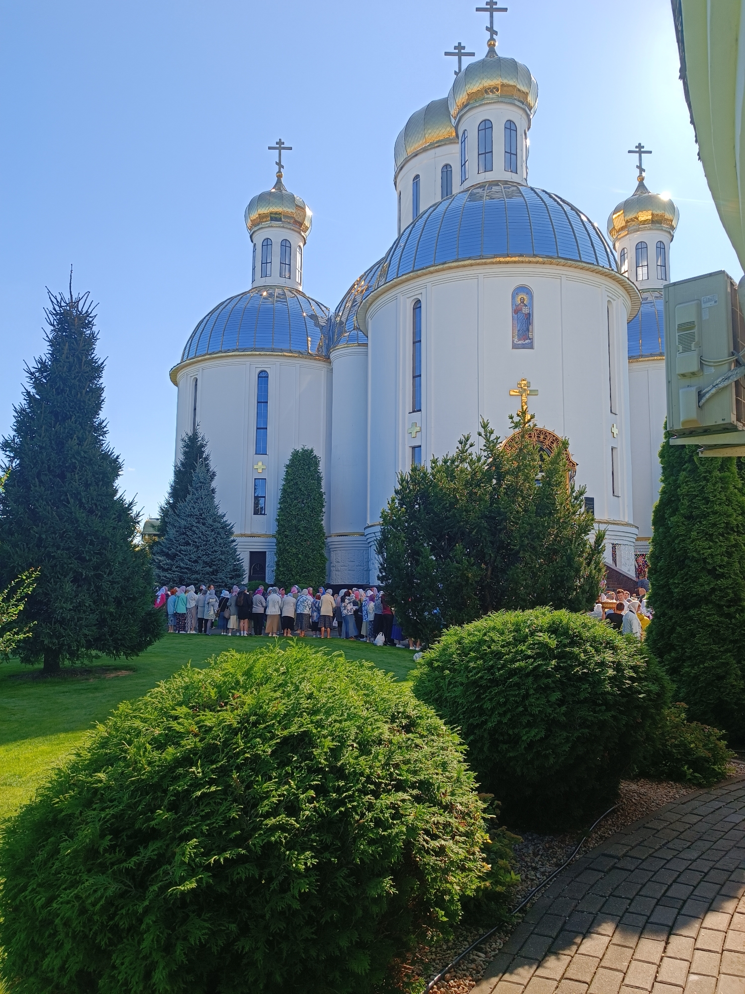 The image shows a group of people standing in front of a white building with a gold-domed roof and blue glass accents, likely a church or cathedral. The building features several crosses and architectural details. There are evergreen trees and manicured bushes in the foreground, with a paved walkway to the right. The sky is clear and blue. The scene appears to be outdoors, on a sunny day, possibly during a religious gathering or event. This is the Holy Assumption Cathedral in Brest, Belarus.