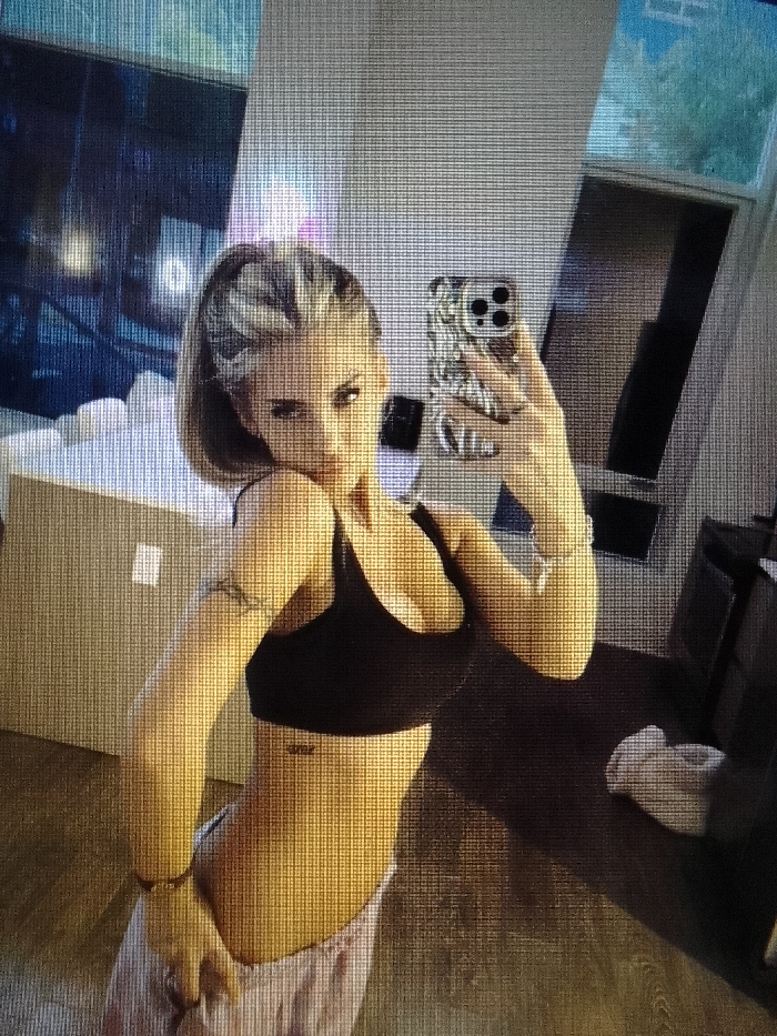 The image depicts a female subject taking a mirror selfie in an indoor residential or studio setting located in the city of Unknown, Unknown. The subject, an adult female with blonde hair pulled into a ponytail, is dressed in a black sports bra and light-colored sweatpants. She holds a smartphone with a distinct patterned case in her right hand, extended to capture her reflection. Her left arm is bent, with the hand resting on her hip area. A small word tattoo is visible on her lower left abdomen, and a faint band-style tattoo is present on her upper left arm.
The environment reflected in the mirror includes a light-colored cardboard box in the lower left, a dark piece of furniture and a crumpled white garment on a wooden floor in the lower right. Reflections of both an exterior night scene with illuminated structures and an exterior day scene with natural light and vegetation are visible through windows in the background. The overall image displays a visible pixelation grid.