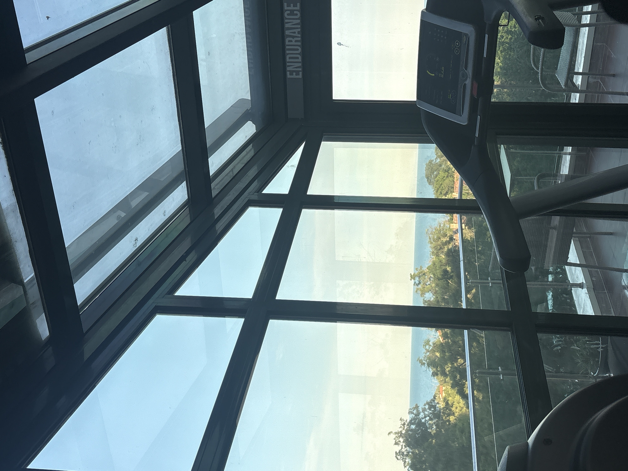 The image shows an interior view of a gym, looking out through a large, multi-paned window. The window frames a scenic view of the ocean and lush green trees. The perspective is from near a treadmill, with its console visible in the upper right portion of the frame. The treadmill's display shows some data, but it's not clearly legible. The window itself is made of glass panes divided by dark frames, creating a grid-like pattern. Some panes are angled, suggesting the room might be a corner space or have a unique architectural design.
Through the window, a calm body of water, likely the sea or ocean, is visible under a bright sky, indicating daytime. The weather appears clear. Dense green foliage, comprising trees and possibly some buildings nestled amongst them, lines the shore. The overall impression is one of a well-lit and modern gym facility offering an attractive view.
On one of the upper window panes, the word "ENDURANCE" is printed vertically in white capital letters against a dark background strip. The lighting suggests it's daytime, perhaps late morning or early afternoon, with the sun illuminating the scene. The setting appears to be a coastal area. There are no people visible in the frame. The mood conveyed is one of tranquility and fitness, with the natural beauty outside complementing the indoor exercise environment.