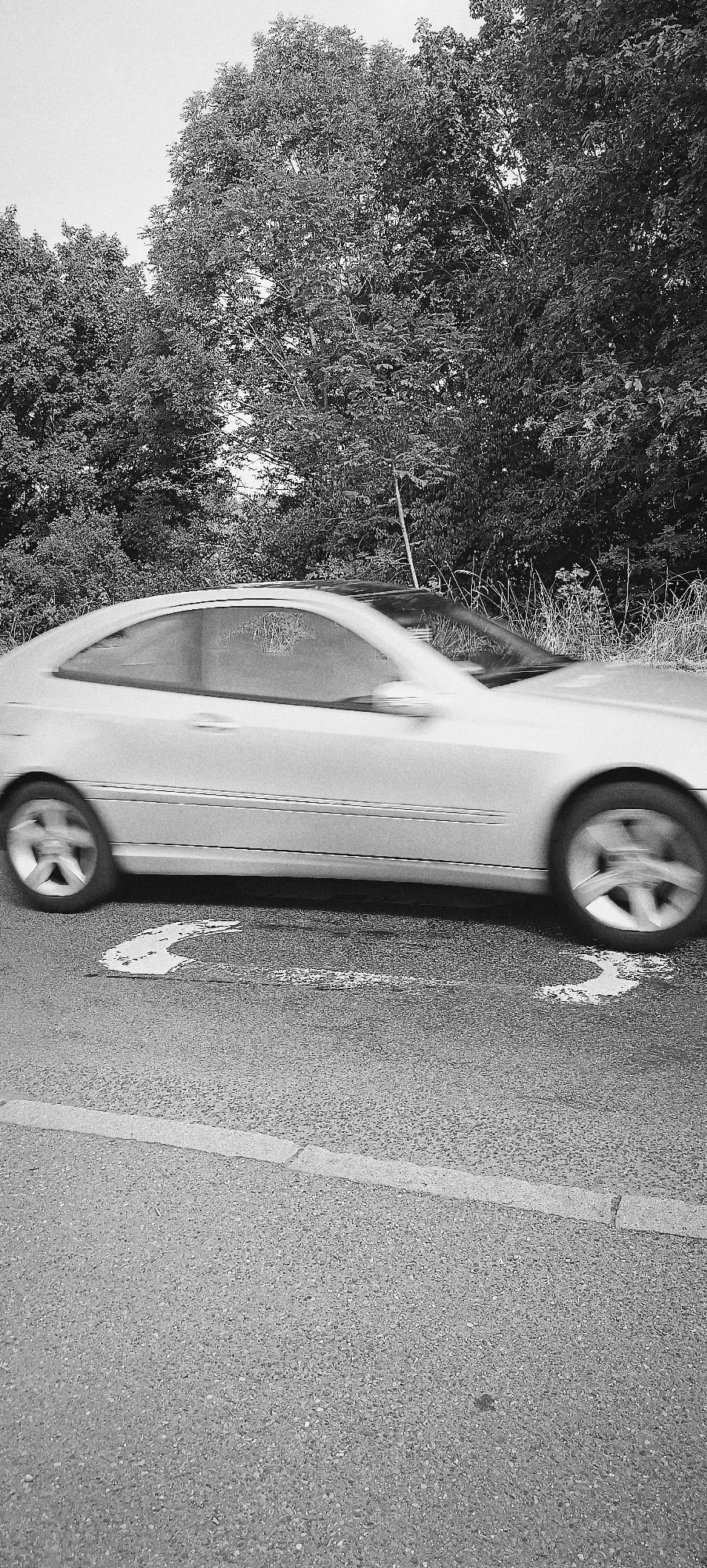 A black and white, medium shot captures a silver coupe driving on a paved road, with a line of trees bordering the right side of the frame. The car is in motion, evidenced by the blur of its wheels and the slight streaking of its body. A white, curved arrow painted on the road indicates a turn or lane change. The trees in the background appear lush and dense, with some grass and weeds growing in front of them. The sky is overcast and washed out. This scene is located on Hauptstraße in Waiblingen, Germany.