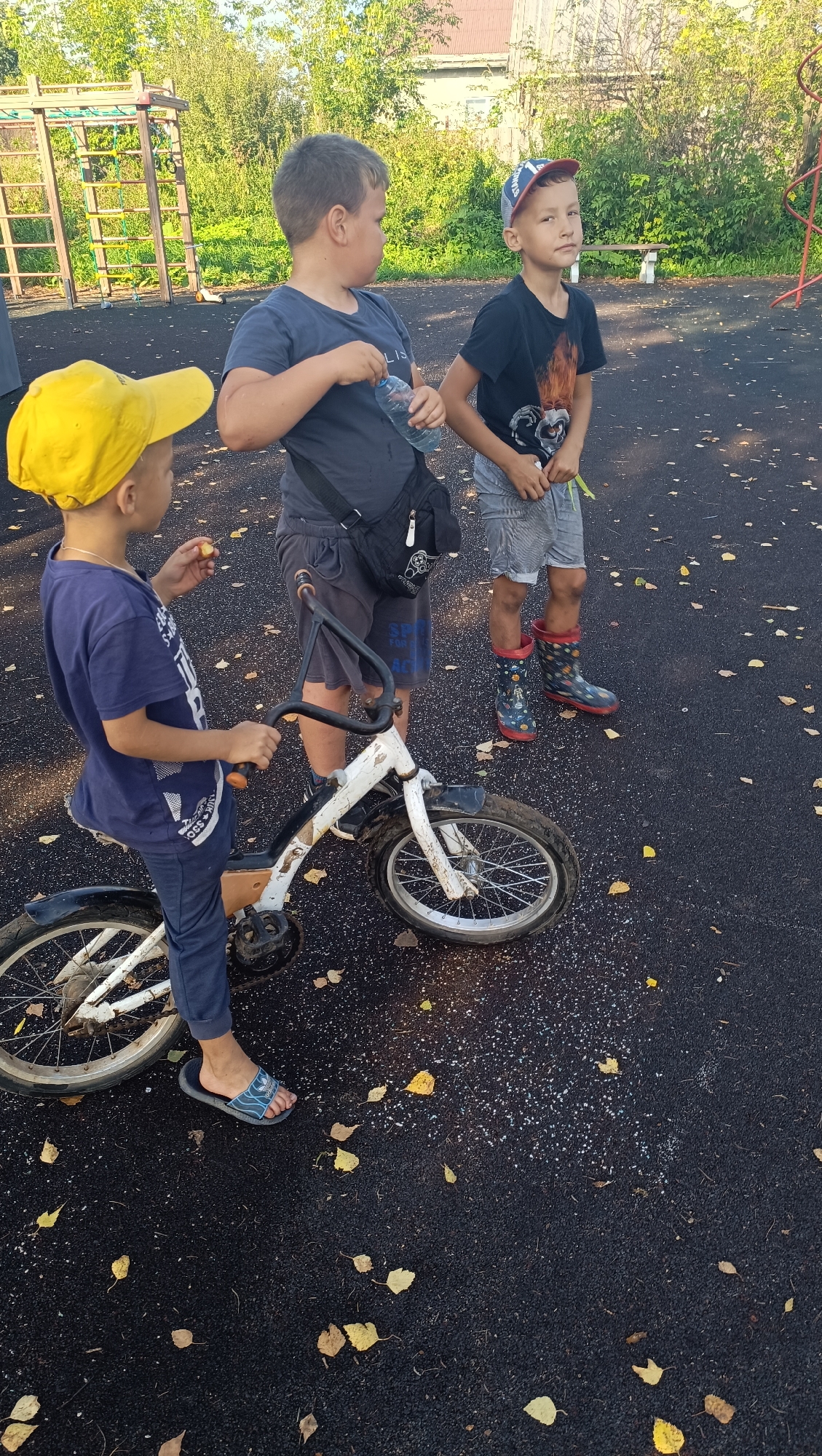In Karsha, Russia, on a playground with a black asphalt surface and scattered yellow leaves, three young boys are present. The boy on the left wears a blue t-shirt, blue pants, and a yellow baseball cap. He is standing next to a small white bicycle, holding a piece of food in his right hand and the bicycle's handlebars with his left. The middle boy, facing left, wears a dark blue t-shirt and dark shorts with a black crossbody bag. He is holding a clear plastic water bottle in his right hand. The boy on the right wears a black t-shirt with a graphic print, gray striped shorts, a red and blue baseball cap, and patterned rain boots. He is looking forward, holding a green object in his right hand. A wooden bench and playground equipment are visible in the background.