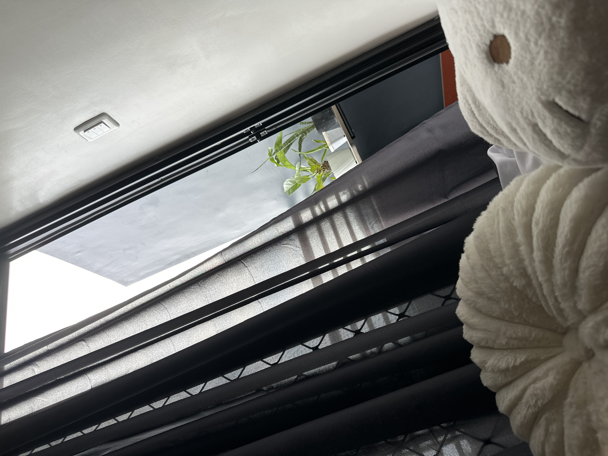 The scene is an interior shot, likely taken in Davao City, Philippines, given the request. The perspective is from inside a room looking outwards through a window covered with semi-transparent black vertical blinds. The window frame is black. Outside, a glimpse of a plant with green leaves is visible, along with part of a grey building and the sky. A white ceiling with a light fixture is seen at the top left corner of the frame. A large, plush, white stuffed animal is partially visible on the right side of the frame, obscuring the view.