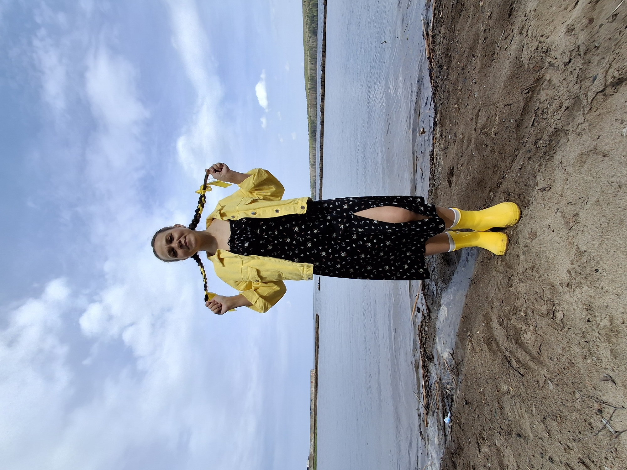 The image shows a young woman standing on a muddy beach. She is wearing a black dress with a floral pattern, a yellow denim jacket, and bright yellow rubber boots. Her hair is braided into two pigtails tied with yellow ribbons. She is holding the ends of her pigtails. The background consists of a body of water, possibly a river or lake, and a cloudy sky. The location is Chaikovsky, Russia.