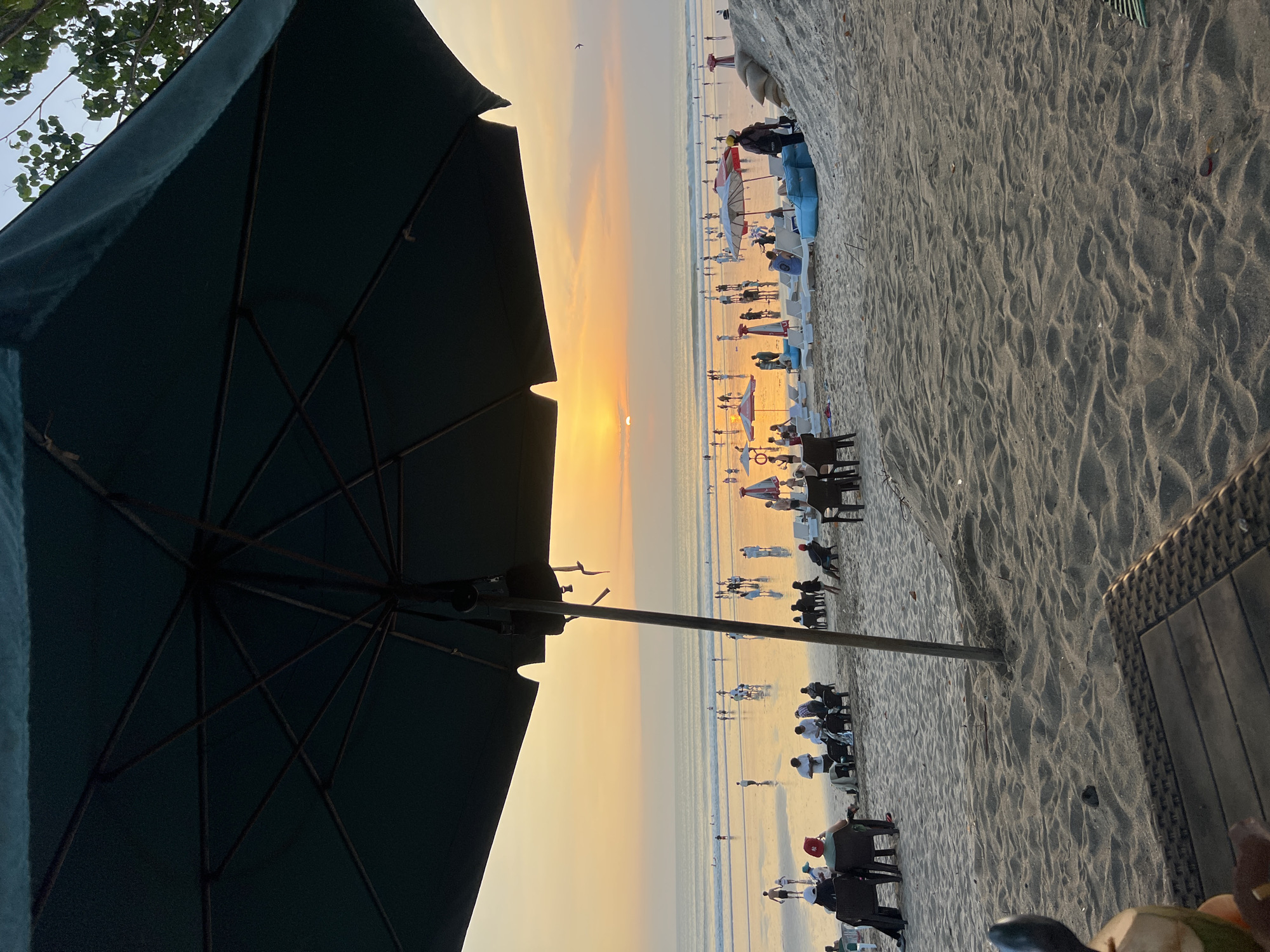 The image depicts a busy beach scene at sunset in Kuta, Indonesia. A large, dark green umbrella dominates the foreground, casting a shadow over a sandy area with a woven table and a halved coconut. Beyond the umbrella, the beach is populated with numerous people, many sitting in chairs or lounging on the sand. Red and white beach umbrellas are scattered across the beach, and several lounge chairs are set up. The ocean is calm, with gentle waves reaching the shore, and the setting sun casts a warm, orange glow across the sky and reflects on the wet sand, creating a shimmering effect. People are seen walking along the shoreline, some wading in the water, and others gathered in groups. The overall atmosphere is relaxed and social, typical of a popular tourist destination at dusk.