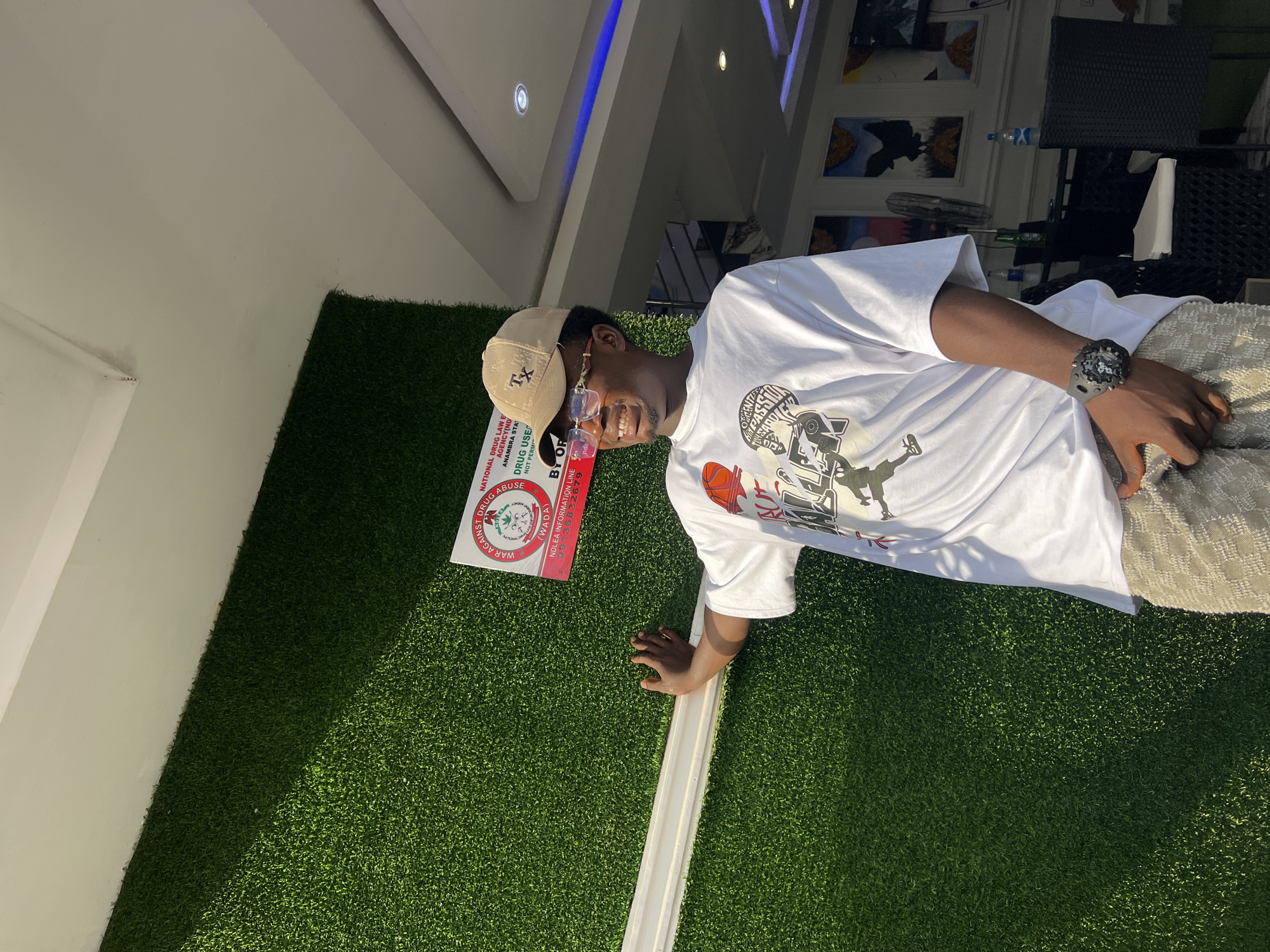 A young Black man wearing a white t-shirt, patterned shorts, a tan baseball cap, and glasses stands with his left hand against a wall covered in artificial grass. He is smiling and looking to his right. A sign with the text "WAR AGAINST DRUG ABUSE" is attached to the artificial grass wall to his left. The man is outdoors with a building interior visible behind him, featuring chairs, tables, and artwork on the walls. The scene appears to be located in Nkpor, Nigeria.