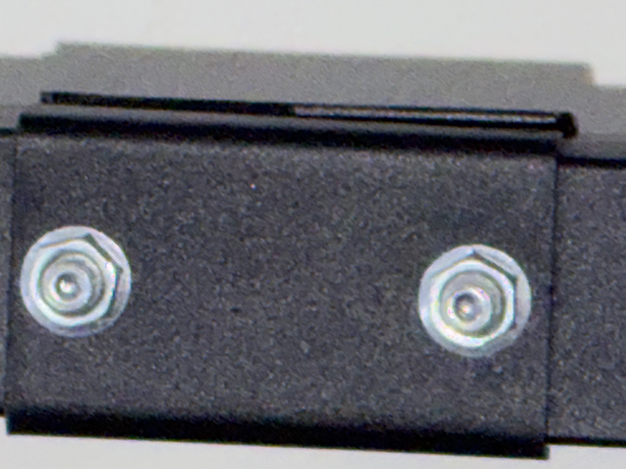 The image is a close-up, vertically oriented photograph of a dark, textured object with two hexagonal nuts visible. The nuts are silver and appear to be part of a mechanical assembly. The object on which the nuts are mounted has a rough, granular surface. To the right of the dark object, there is a thinner, darker vertical element. The background is a light, neutral color, suggesting an indoor setting or a controlled environment. The image focuses on the details of the hardware, highlighting the texture of the mounting surface and the reflective properties of the nuts. The location is not discernible from the visual information provided in the image.