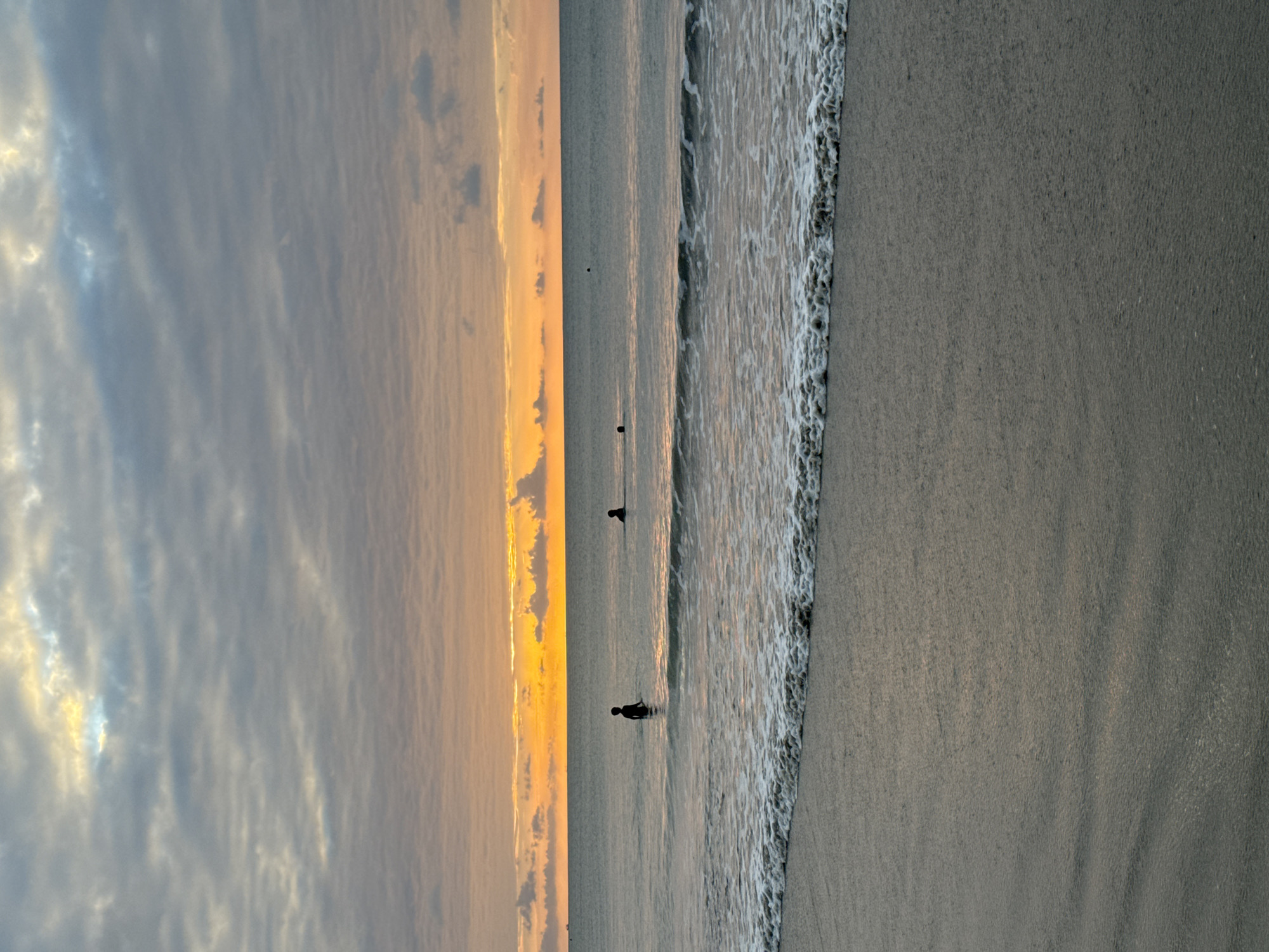 An outdoor, waist-level shot captures a serene beach scene at sunset in Kuta Selatan, Indonesia. The sky is a gradient of soft orange and yellow near the horizon, transitioning to a muted gray with wispy clouds overhead. The ocean is calm and appears grayish-blue, with gentle waves lapping at the shore. The foreground is dominated by a wet, sandy beach reflecting the muted colors of the sky. Three human figures are present in the water: one standing silhouetted in the middle ground, and two others further out, also appearing as dark silhouettes against the water and sky. The overall atmosphere is tranquil and peaceful, typical of a sunset on a tropical beach.