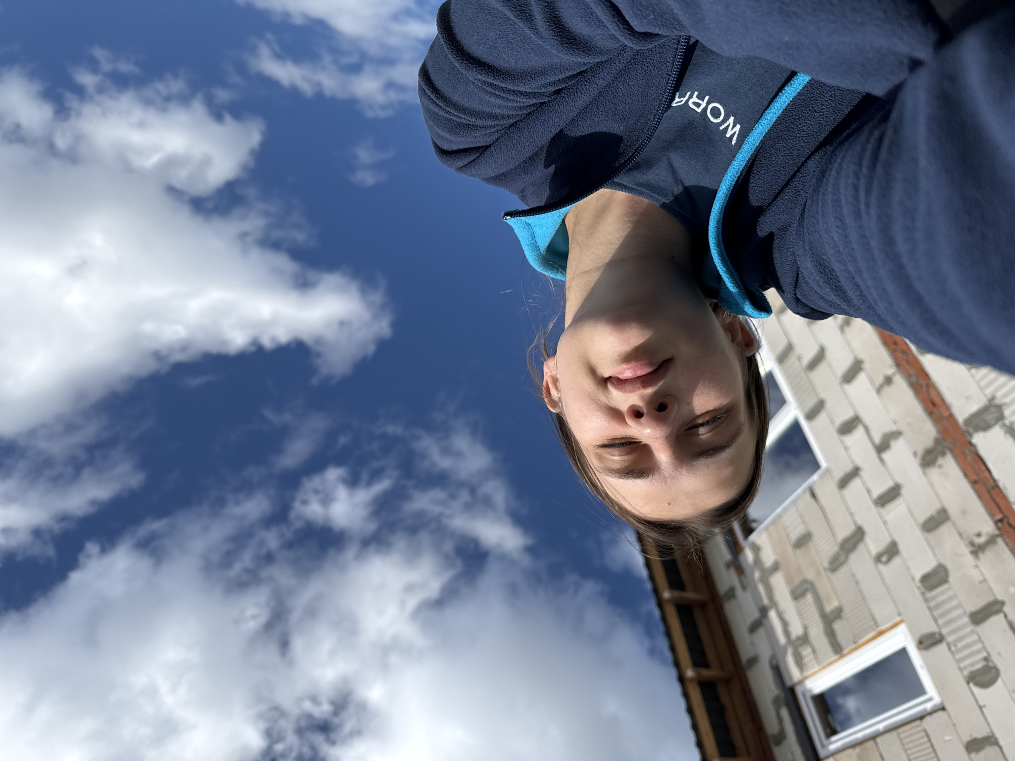 This is a selfie taken from a low angle, looking up at the sky and the subject. The subject is a young person wearing a navy blue fleece jacket with a bright blue trim around the collar. The jacket has the word "WORK" printed on the chest in white letters. The person's eyes are closed, and they have a slight smile, suggesting a relaxed or peaceful emotion.
The background is dominated by a bright blue sky with scattered white clouds. To the right of the subject, a building under construction is visible. It appears to be made of pale concrete blocks with some reddish-brown brick accents. There are at least two windows visible on the building, one with a white frame and the other with a darker frame, both reflecting the sky. The overall scene suggests an outdoor setting on a clear day. The time of day appears to be daytime, likely midday or afternoon, given the brightness of the sun and sky. The location context provided is Chaikovsky, Russia.