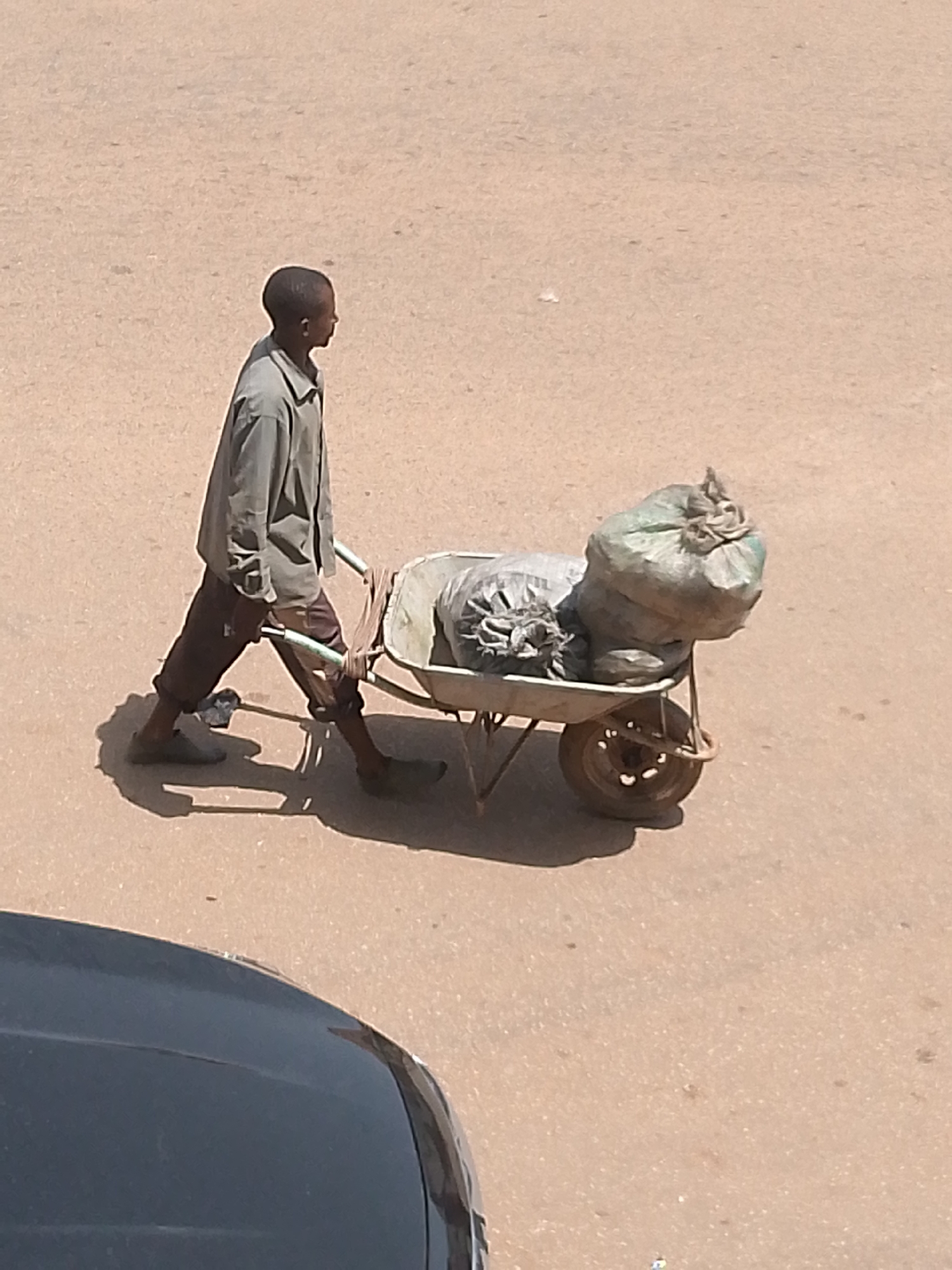 An underexposed, eye-level shot captures a young boy from a medium distance, pushing a wheelbarrow filled with two large sacks on a paved road. The boy, seen from the side, wears a loose-fitting, light-colored long-sleeved shirt and dark trousers. His left hand grips the wheelbarrow's handle, while his right hand is further back. The wheelbarrow is metal with a single wheel and two handles. The sacks in the wheelbarrow appear to be made of a dark material, one larger and more rounded than the other. The road surface is a neutral tan color, and a dark car hood is partially visible in the lower left corner, casting a shadow on the road. The lighting suggests it is daytime.