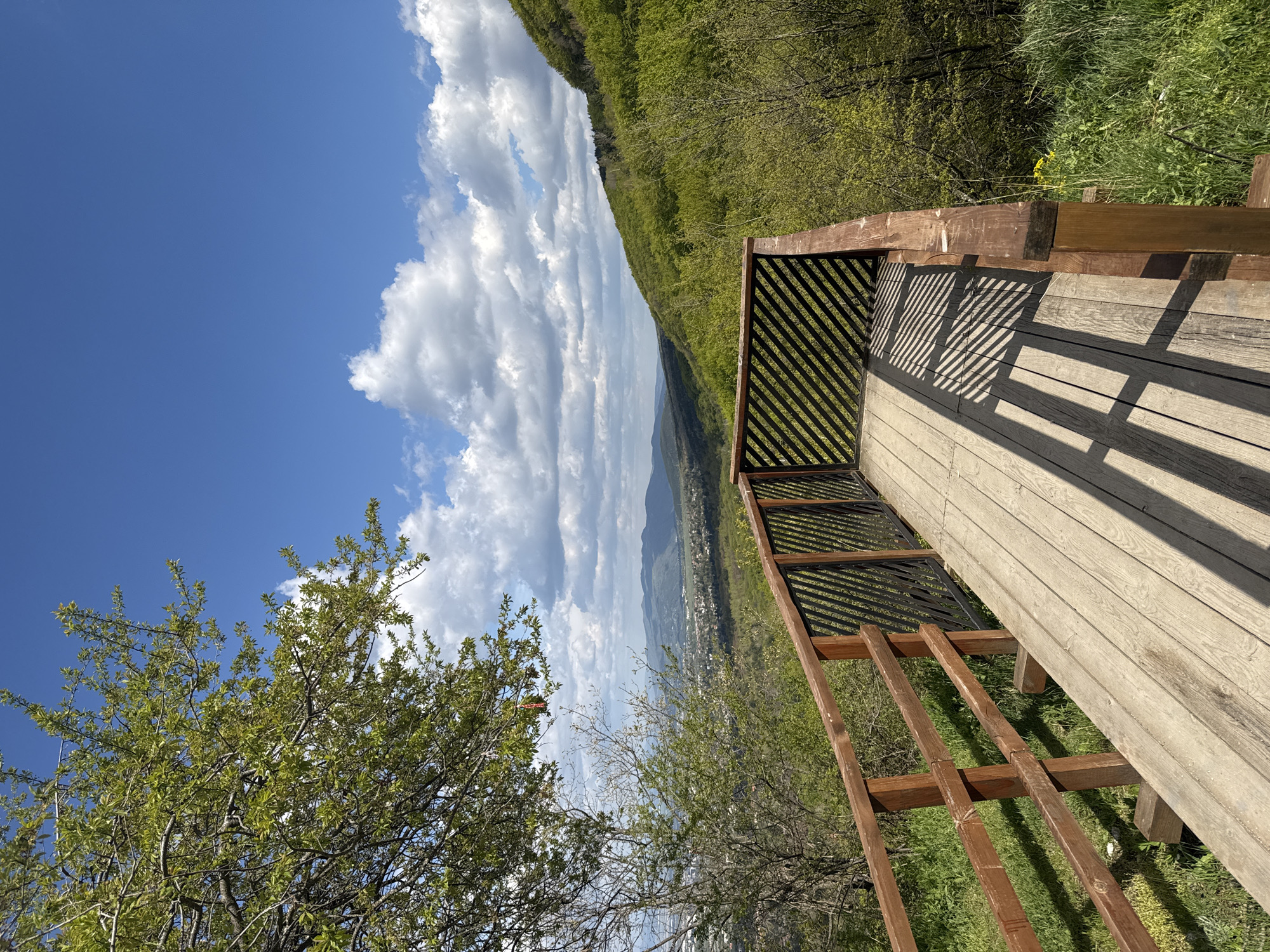 A horizontal, medium-distance shot depicts a wooden platform overlooking a scenic landscape in what appears to be a mountainous region on a sunny day with puffy white clouds in Sofia, Bulgaria. The platform is made of light-colored wood planks with dark-colored wooden railings, creating striped shadows on the surface. Behind the platform is a lush, green forested slope, receding into distant mountains shrouded in a slight haze. Below the mountains lies a town or city. A green-leaved tree occupies the bottom left corner of the frame, contrasting with the deep blue sky and clouds.