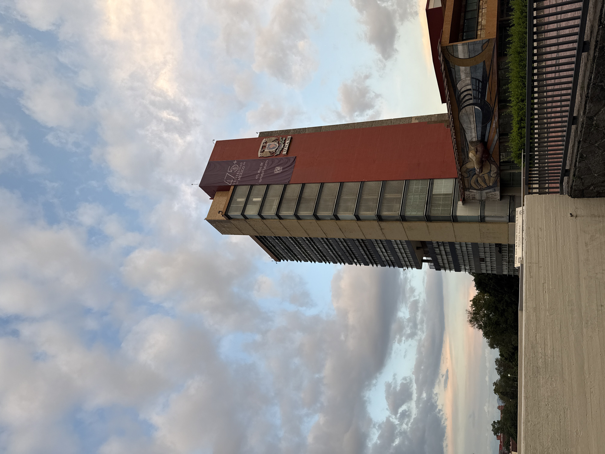This is a medium shot of the UNAM Central University City campus in Mexico City. The dominant subject is a tall, multi-story building with a red facade and a tall, narrower section made of glass and beige brick. A large banner hangs from the red section of the building, celebrating "475+ Años de construir futuros" and featuring the UNAM emblem and logo. Another building with a mural is visible to the right, featuring a depiction of a hand holding a cylindrical object. A concrete wall and a black metal fence are in the foreground. The sky above is partly cloudy with hues of blue, white, and pink, suggesting either early morning or late afternoon light. Trees are visible in the background.