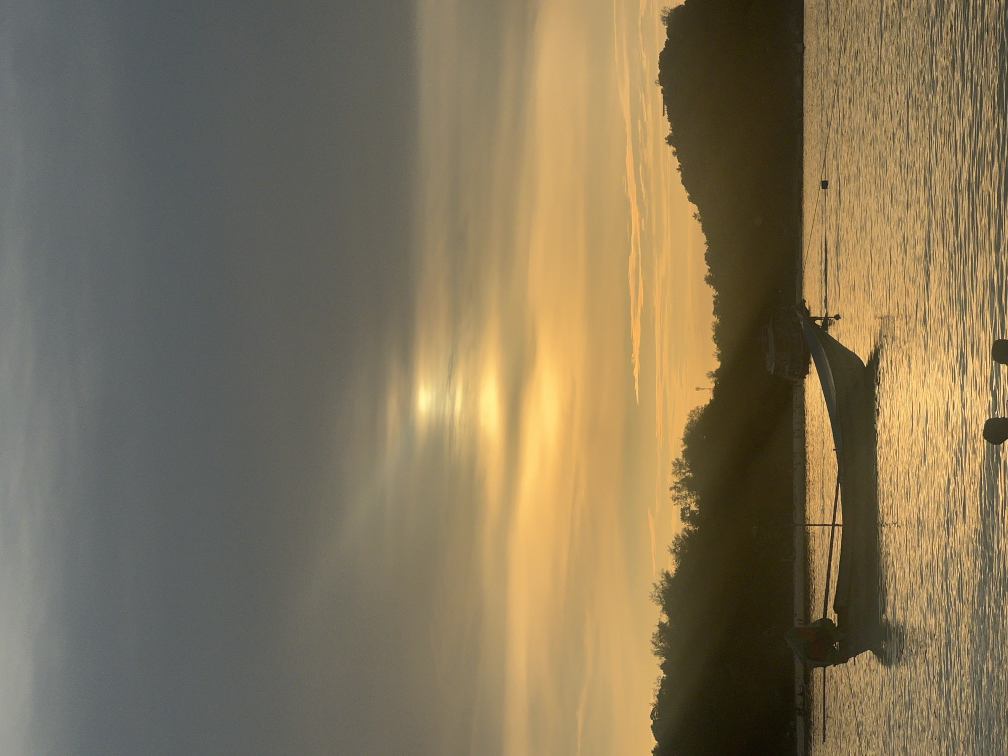 A vertical, eye-level, medium shot captures a serene sunset over a body of water, likely the sea, in Ko Pha-ngan District, Thailand. The sky transitions from a deep gray at the top to a vibrant golden orange near the horizon, with thin, elongated clouds streaking across the upper portion. The sun, a bright orb, is partially obscured by clouds, casting long, dramatic rays of light that pierce through the atmosphere. In the middle ground, a silhouetted, dark green treeline of hills forms the coastline.
Dominating the foreground is the calm, shimmering surface of the water, reflecting the warm hues of the sunset. A solitary, traditional wooden fishing boat, with a pointed bow and a simple canopy structure, is moored in the water, its silhouette also dark against the bright reflections. The boat appears to be empty, with a single oar resting across its width. Further in the background, another larger, silhouetted boat is visible on the water. Two dark, indistinct figures, possibly people, are visible in silhouette at the very bottom of the frame, their forms partially submerged in the water. The overall impression is one of tranquility and natural beauty.