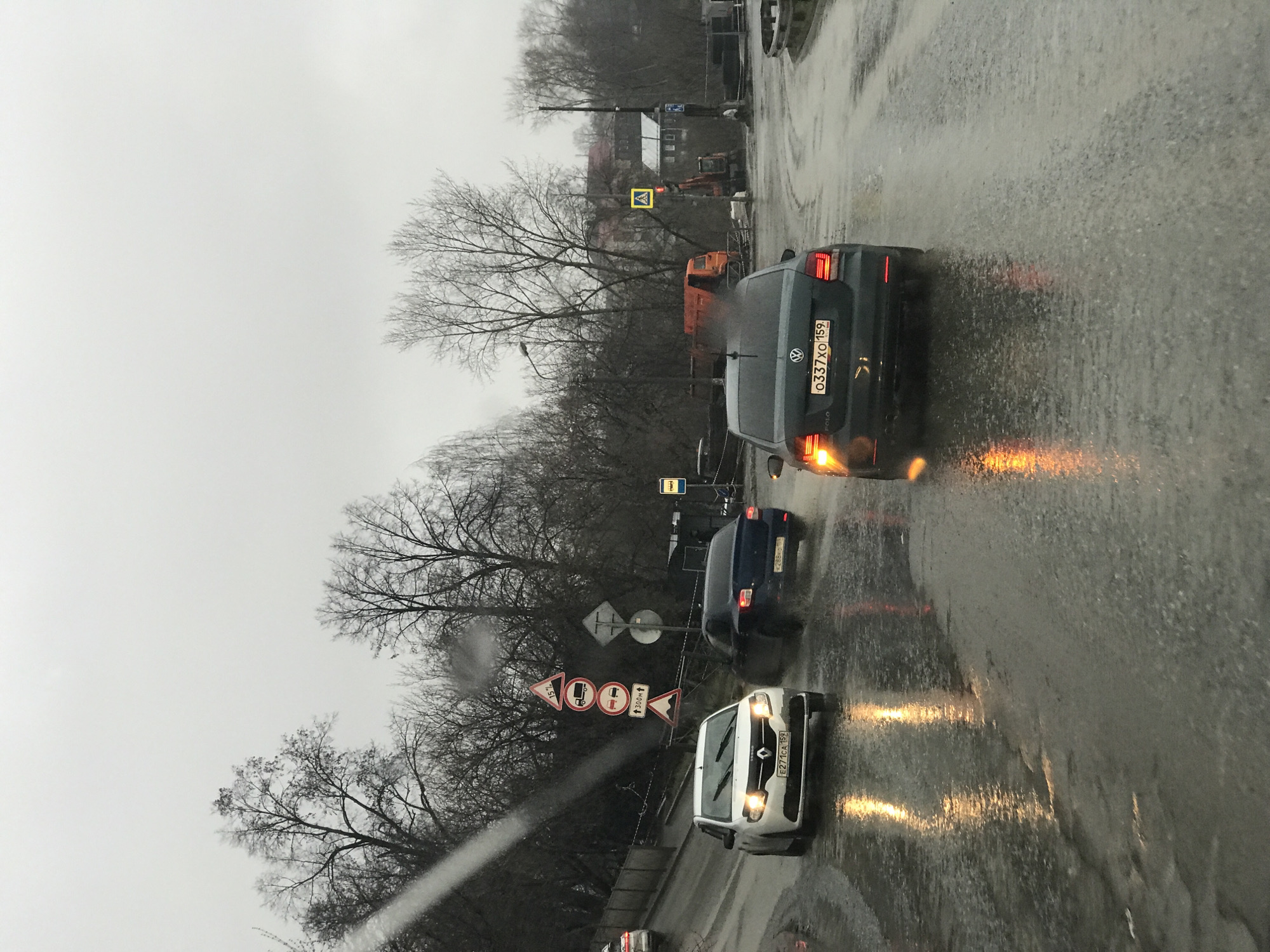 The image depicts a rainy street scene in Perm, Russia. Traffic is visible, consisting of several cars: a dark gray Volkswagen Polo with license plate "O337 XO 159", a blue car, and a white Renault Kangoo with license plate "E271 CA 159". Road signs are present, including a pedestrian crossing sign and signs indicating road restrictions, such as height, weight, and length limits for vehicles, as well as a sign indicating a distance of 300 meters. Trees without leaves line the road. In the background, buildings and construction equipment are visible. The road surface is wet and reflecting the lights of the vehicles.