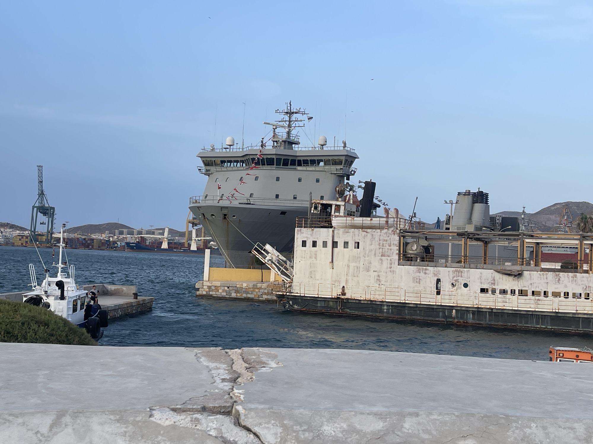 In Cartagena, Spain, a large grey naval vessel is docked alongside a derelict white ship. The naval vessel has several antennas and navigation equipment on its bridge, and small flags are flying from its superstructure. The white ship appears weathered and rusted, with visible signs of decay. In the foreground, a small white tugboat is moored to a stone jetty where two individuals are standing. A rough concrete pier occupies the immediate foreground. In the background, cargo ships and cranes are visible in the harbor, indicating an active port. The sky is clear and light blue.