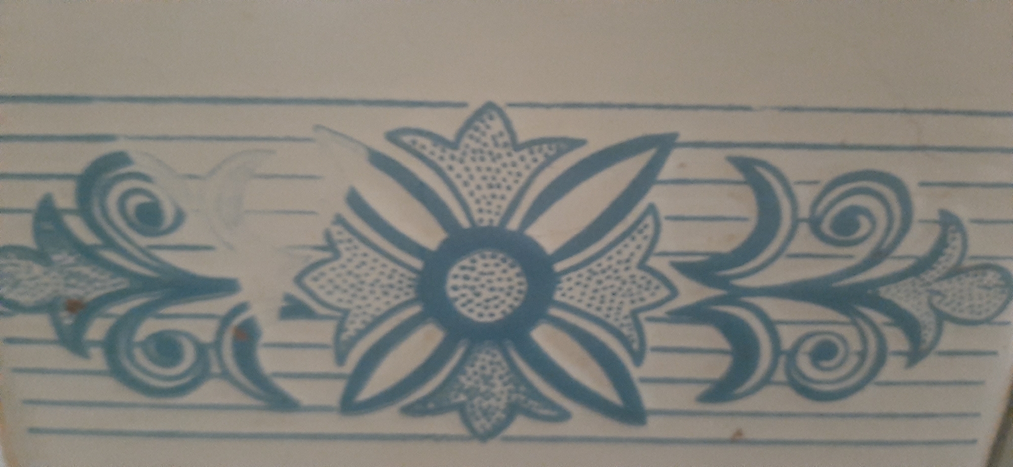 The image displays a close-up of a decorative border pattern on a light-colored surface, possibly ceramic tile or wallpaper. The pattern features stylized floral motifs in a medium blue color, set against a background of thin, parallel blue lines that run horizontally. The central element is a large, symmetrical flower-like design with layered petals filled with stippling. Flanking this central motif are swirling, abstract shapes that resemble foliage and scrolls, also outlined in blue and some areas filled with stippling. There are a few small, reddish-brown spots scattered across the surface, which may be imperfections or part of the material. The lighting appears to be even, casting no strong shadows, suggesting an indoor setting or diffused light. The image quality is somewhat blurry and low resolution, making fine details difficult to discern. There are no people, animals, or any indication of a specific location within Yessentukskaya, Russia. The image solely captures a decorative pattern.