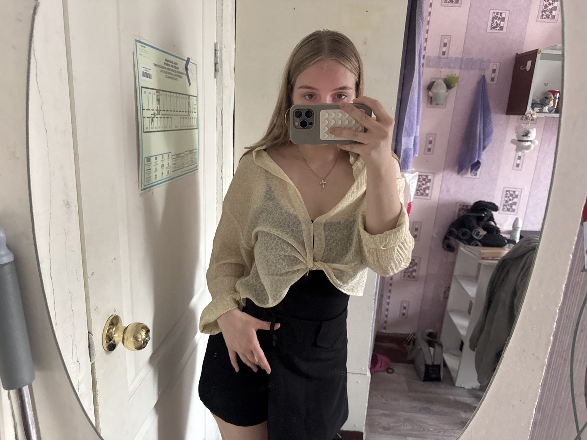 A young woman is taking a mirror selfie in what appears to be a room in an apartment building. She is wearing a sheer, light-colored button-up shirt tied at the waist over a black top and a black skirt. She has blonde hair and is holding a phone with a PopSocket in her right hand. The room has pale pink striped wallpaper with a pattern of squares, and various items are visible in the background reflected in the mirror, including shelves with objects, towels, and what appears to be a mop. A door with a brass doorknob is to her left, and a notice is affixed to it. The overall impression is of a private living space. The city of Perm, Russia is not directly discernible from the image, but the style of the wallpaper and general decor might suggest a setting common in Russian residential areas.