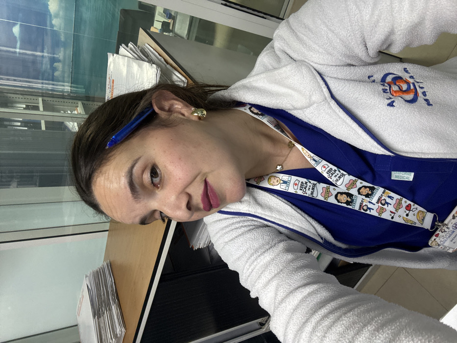 A woman is captured in a selfie-style photograph taken indoors in Meknes, Morocco, showing her head and upper torso. Her dark hair is pulled back, with a blue pen tucked behind her right ear. She wears small gold earrings and a delicate gold necklace with a four-leaf clover pendant. Her lips are adorned with a muted berry lipstick, and she maintains a neutral to subtly pleasant expression while looking towards the camera.
She is dressed in a royal blue scrub top, visible beneath an open white fleece jacket. The jacket features an embroidered logo on the right chest that reads "Alma Mater." Around her neck hangs a white lanyard, decorated with various colorful cartoon illustrations and text. Prominently visible phrases on the lanyard include "KEEP CALM I'M A NURSE" and "DANCE IT OUT." A tag on her scrub top clearly states "UNIFORME MEDICAL."
The background shows an office-like setting with a large window revealing a cloudy sky. A stack of papers or documents is visible on a light-colored wooden desk or counter, suggesting an administrative area within what appears to be a medical facility. The image is rotated, presenting the scene from an oblique angle.
