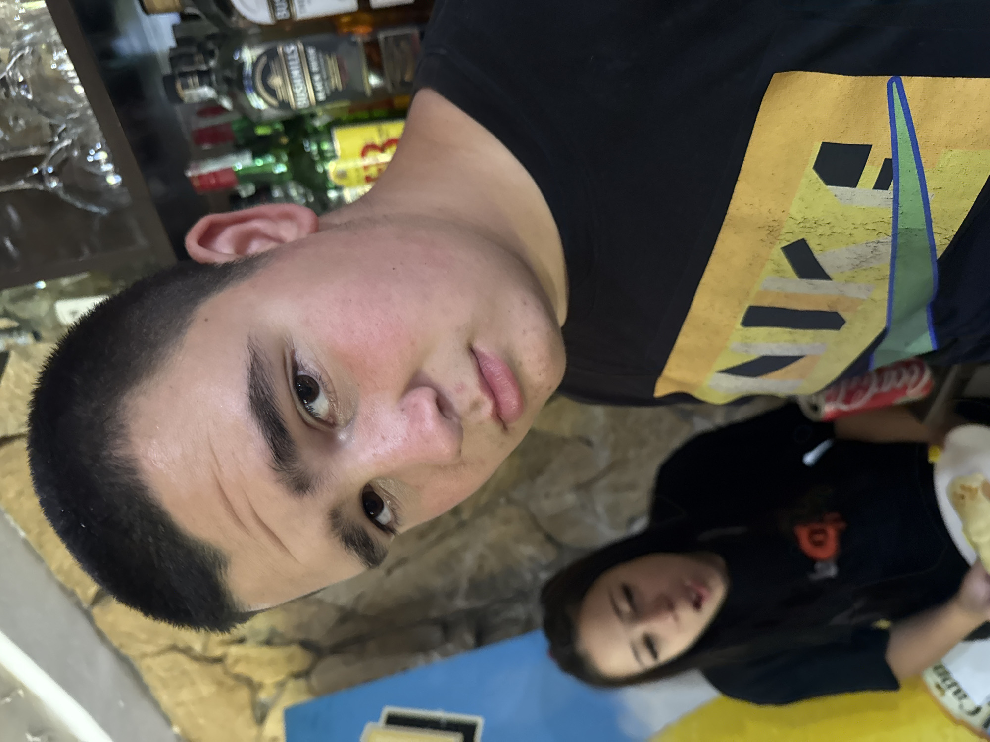 A young man with a short haircut is looking directly at the camera, his expression neutral. He is wearing a black t-shirt with a yellow and multi-colored graphic design. Behind him, to his left, a young woman with dark hair is blurred and appears to be speaking or eating, holding a plate with food. She is wearing a black t-shirt. In the background, shelves with various bottles and glassware suggest a bar or liquor store setting. The wall behind the subjects is textured and appears to be made of stone or rough plaster. A bright blue sign with yellow lettering is partially visible on the left side of the frame. A red and white can, likely a soda, is visible next to the woman. The lighting is moderate, with some shadows. The image is taken from a slightly low angle. The location in Sredets, Bulgaria is not discernible from the visual information provided.