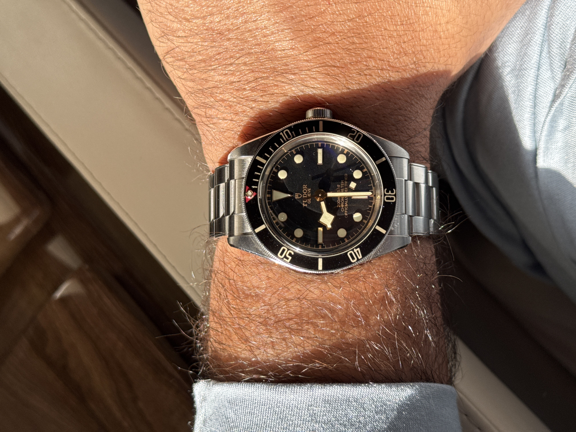 A close-up shot features a Tudor Black Bay Fifty-Eight wristwatch on a man's wrist. The watch has a black dial with gold accents, a stainless steel case, and a matching bracelet. The watch is positioned on the man's left wrist, which is covered in hair. The man wears a light blue collared shirt. The background shows a wooden surface and a light-colored cushion or seat. The location in the city of Dubai, United Arab Emirates cannot be determined from this image.