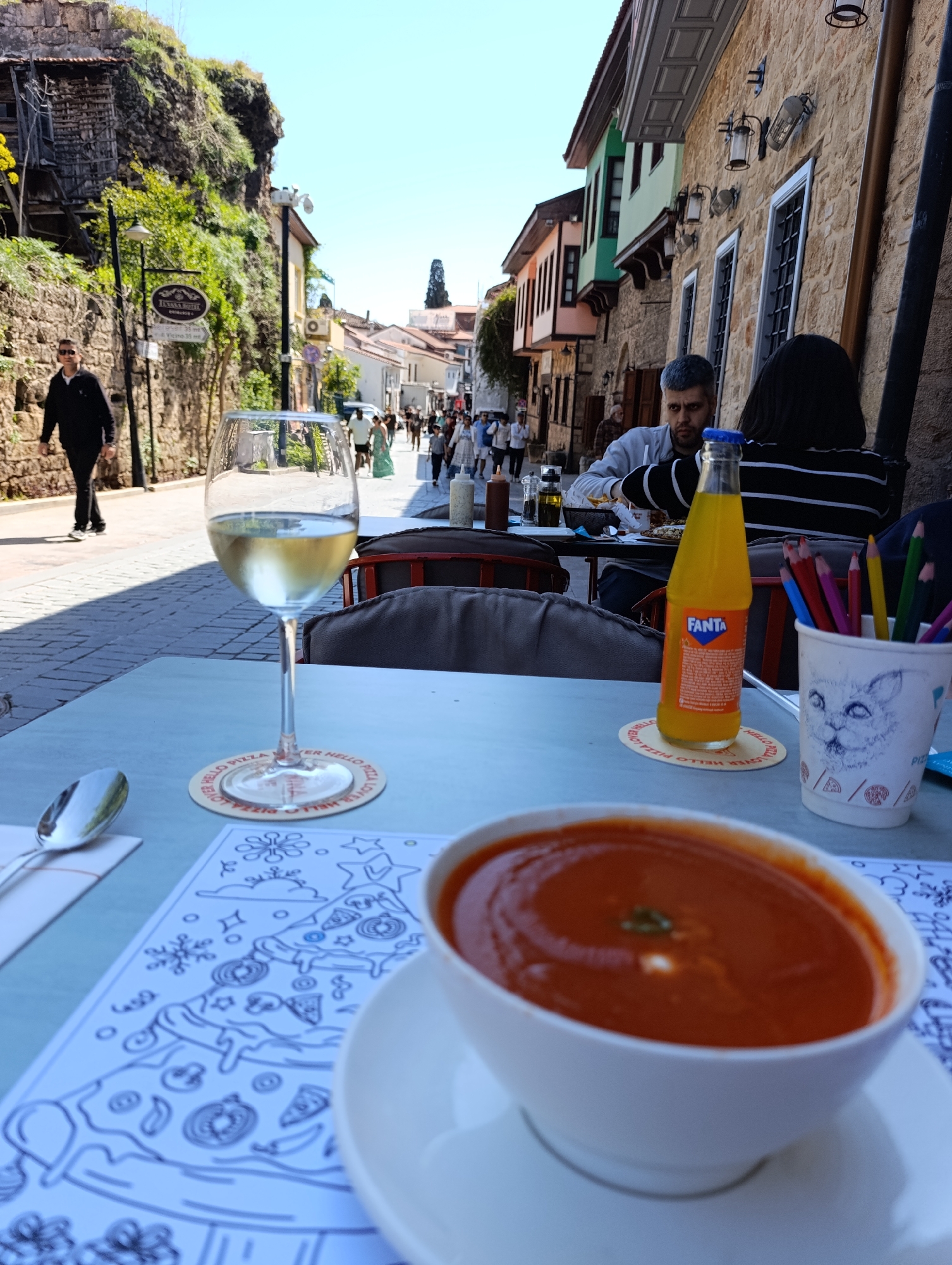 A street in Antalya, Turkey, is viewed from a table in an outdoor dining area. The foreground features a bowl of tomato soup on a saucer, a coloring page with pizza designs, a spoon on a napkin, a glass of white wine, a bottle of Fanta orange soda, and a paper cup filled with colored pencils, set on a blue-grey table. In the midground, a man sits at a table, eating. Other people stroll down the paved street. Buildings line both sides of the street, constructed with stone and featuring architectural details. The sky is clear and blue. A tree-covered hillside rises on the left side of the street.