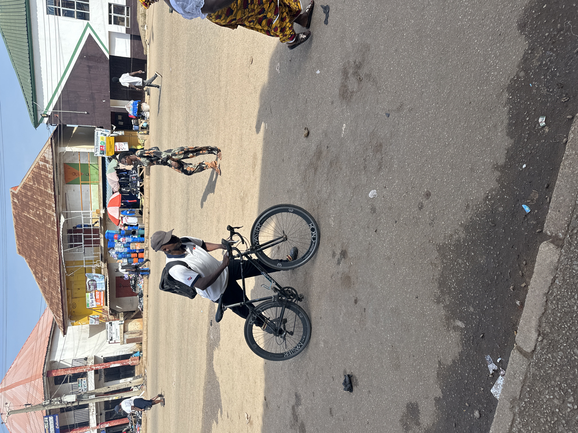 The image shows a street scene in Bukuru, Nigeria. In the foreground, a man wearing a white polo shirt, black trousers, and a baseball cap is leaning over a black bicycle. He has a backpack on. The bicycle is a road bike with deep-section rims. Behind him, several other people are walking or standing. To the left, a woman in a white t-shirt and dark shorts is walking. Further back, buildings line the street, with stalls and various goods displayed in front of them. There are also stacks of blue plastic containers. The ground is asphalt and appears somewhat dusty and worn. The sky is a clear blue.
