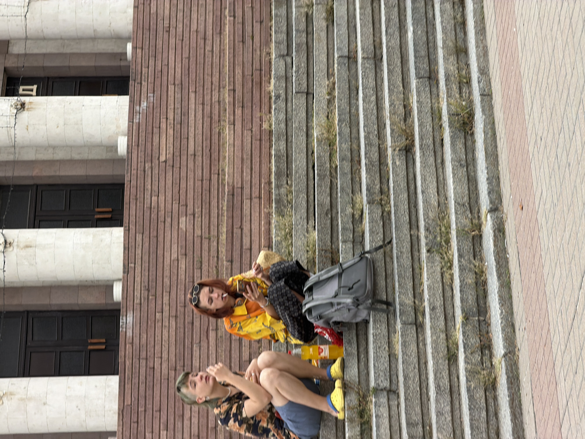 Two individuals are seated on stone steps outside a building with large classical columns and dark double doors. The person on the left, with short green-tinted hair and wearing a patterned short-sleeved shirt and blue shorts, is looking away from the camera and appears to be smoking or holding something to their mouth. They are wearing yellow clogs. The person on the right, with red hair and wearing sunglasses on their head, is looking at their phone and smoking. They are dressed in a brightly colored yellow and orange patterned shirt and black patterned pants. Beside them is a bottle of yellow liquid, a red bag, and a grey backpack. The steps are made of large grey stones and the background features red stone steps leading up to the building. The scene appears to be outdoors in a public space. The location is identified as Kyiv, Ukraine.