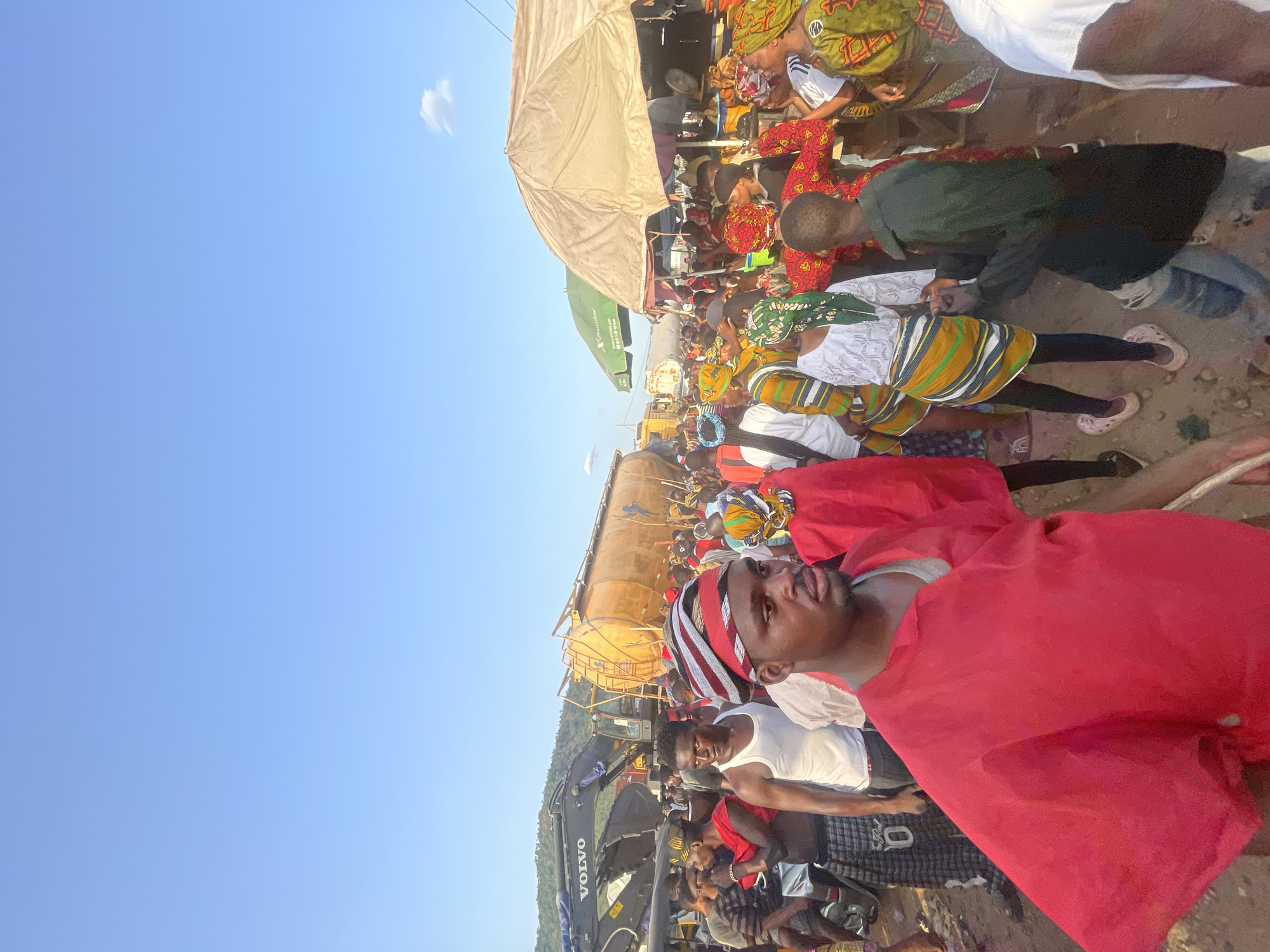 The image depicts a scene in Lokoja, Nigeria, crowded with people gathered around a large tanker truck. A man wearing a red cloth and a striped head covering is visible in the foreground. The tanker appears to be the focus of attention, with individuals standing around and on top of it. Some people are wearing traditional African clothing and head wraps. An excavator and a Volvo truck are also present, suggesting ongoing construction or activity in the area.